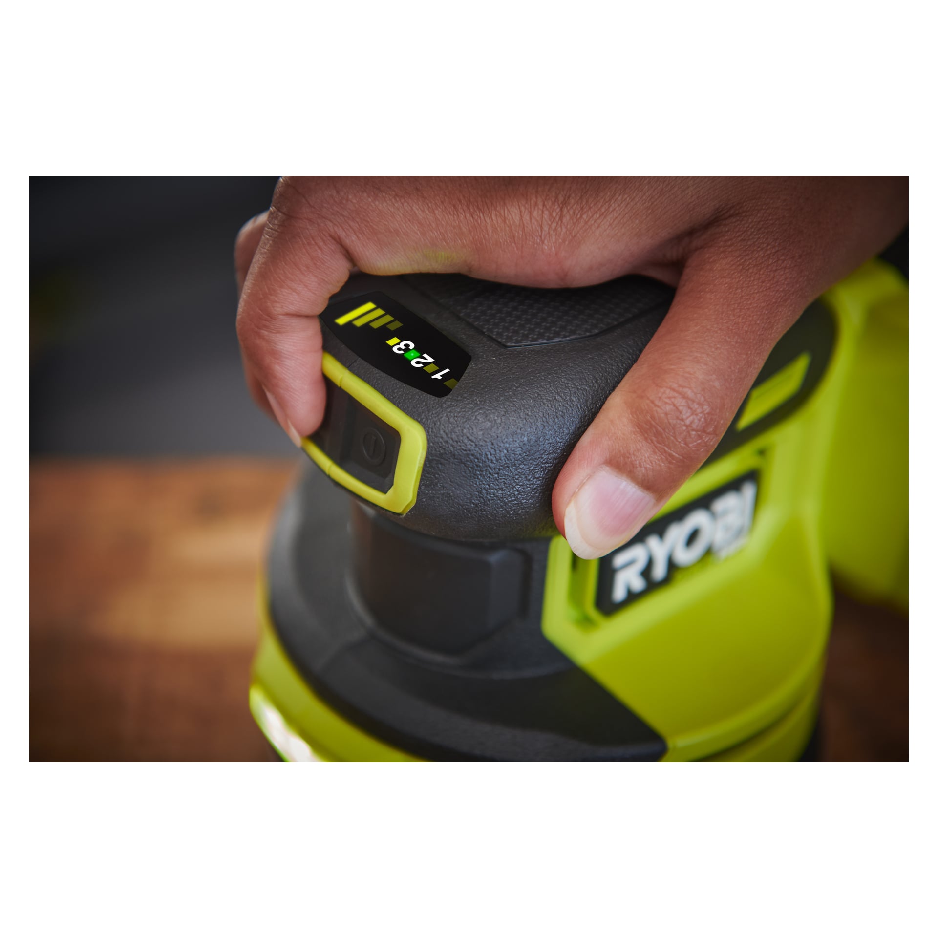 Eine detaillierte Ansicht einer Hand, die einen Ryobi One+ HP Eccentric Sander RROS18C-0 hält, Daumen drückt einen Knopf neben einem digitalen Bildschirm mit 12 2 3, mit einem Teil des Ryobi One+ HP Logos im Hintergrund.