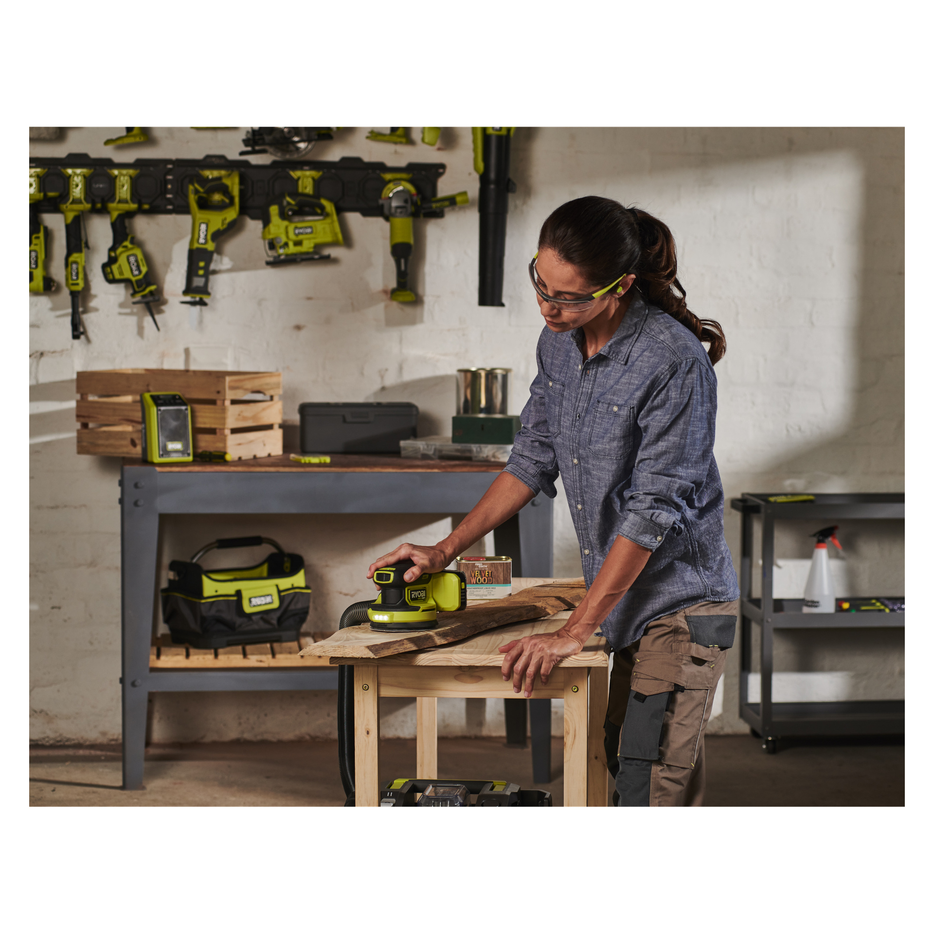 Eine Person mit Sicherheitsbrille schleift ein Holzbrett mit dem Ryobi One+ HP Eccentric Schleifer RROS18C-0 in einer gut organisierten Werkstatt, umgeben von Regalen und Regalen mit verschiedenen Werkzeugen und Ausrüstung.