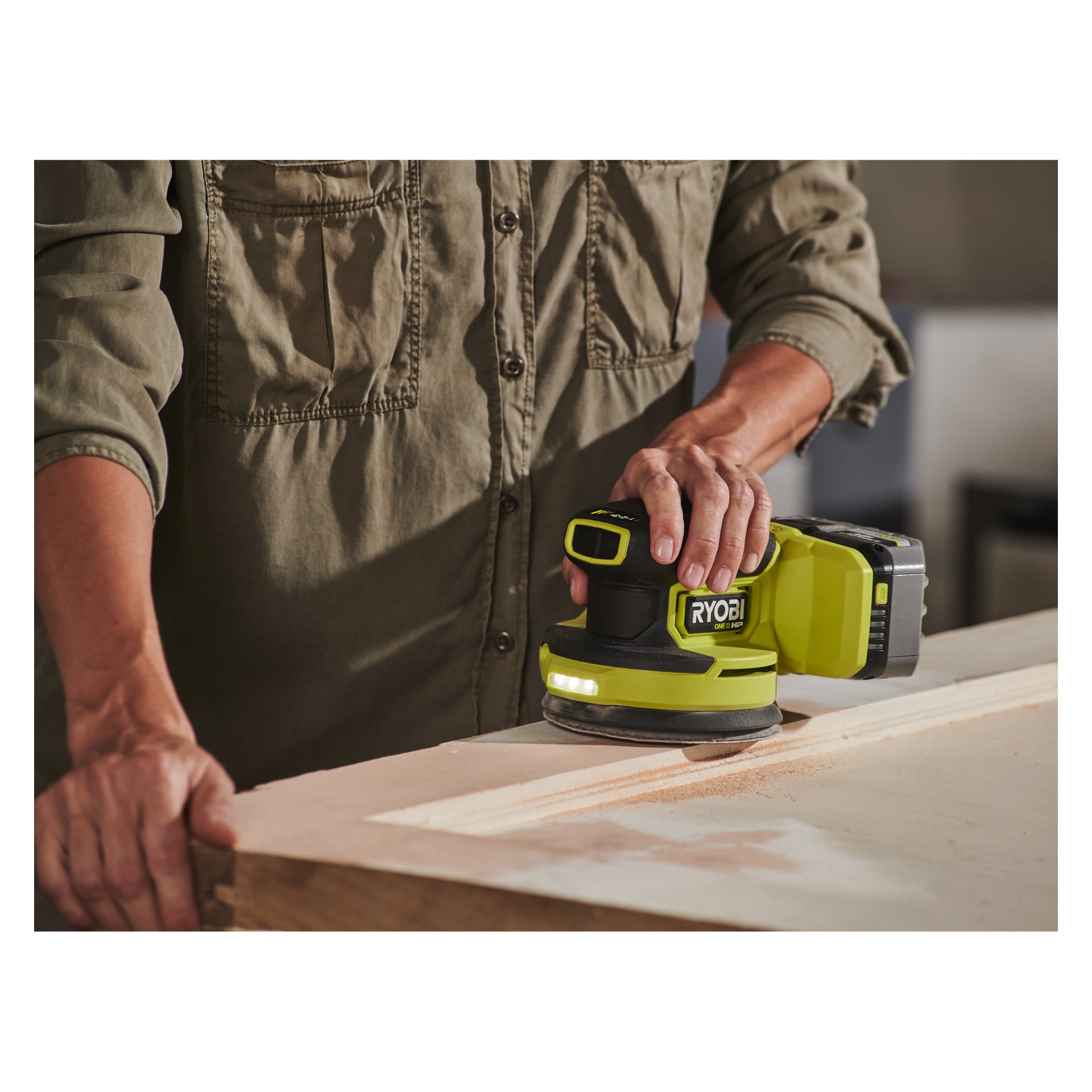 Eine Person in einem grünen Hemd bedient einen Ryobi One+ HP Eccentric Sander RROS18C-0 auf einer Holzoberfläche, wobei beim Glätten des Holzes Sägemehl entsteht.