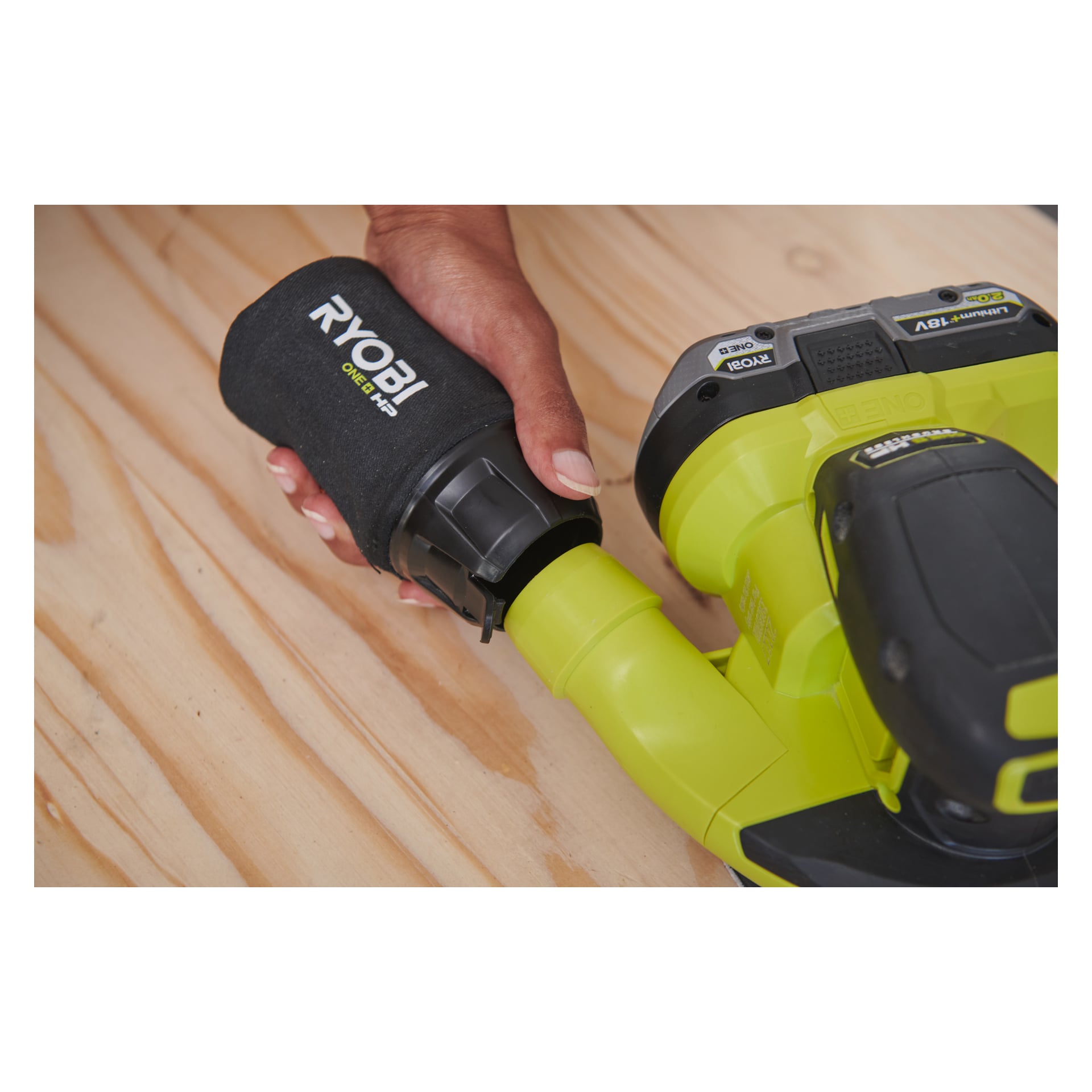 Eine Hand verbindet einen schwarzen Staubsack mit der Aufschrift Ryobi One+ HP an den Staubport eines grünen RROS18C-0 Exzentrischen Schleifers, der auf einer leichten Holzoberfläche ruht. Die Branding- und Steuerungseinstellungen des kabellosen Werkzeugs sind nahe dem Griff sichtbar.