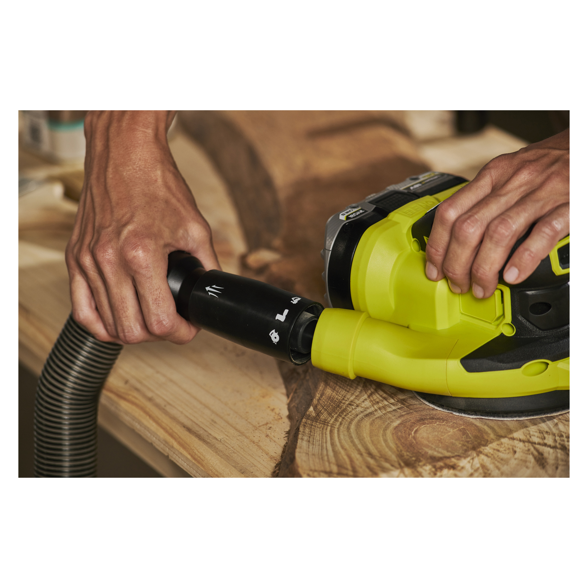 Zwei Hände verbinden einen schwarzen Vakuumschlauch mit einem grünen Ryobi One+ HP Eccentric Schleifer RROS18C-0, der auf einer Holzoberfläche mit markanter Holzmaserung ruht. Der Schwerpunkt liegt darauf, wo Werkzeuge und Hände zusammentreffen.