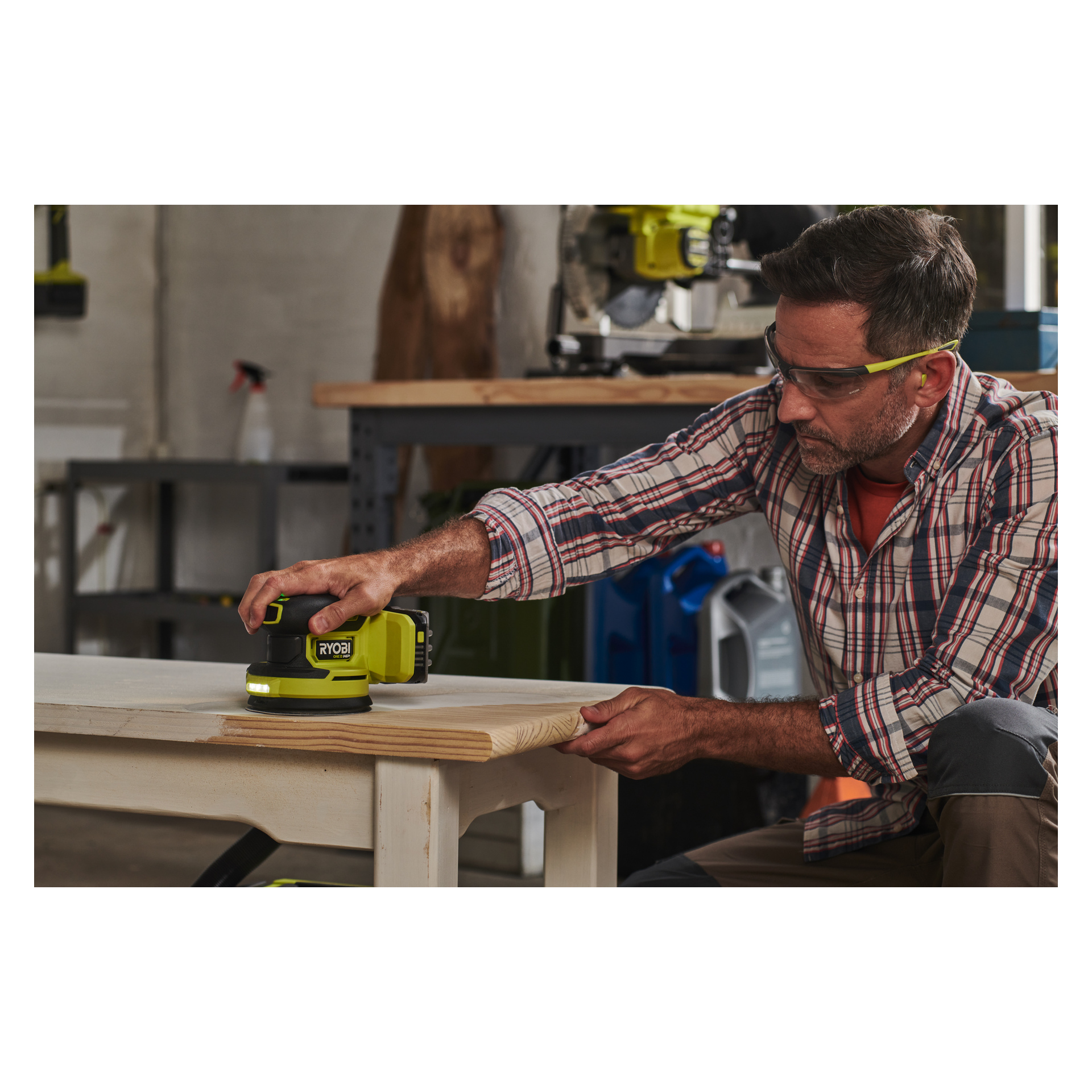 Ein Mann mit Schutzbrille bedient einen Ryobi One+ HP Eccentric Sander RROS18C-0 auf der Ecke eines Holztisches in einer Werkstatt, mit verschiedenen Werkzeugen und Ausrüstung auf einer Werkbank und Regalen hinter sich.