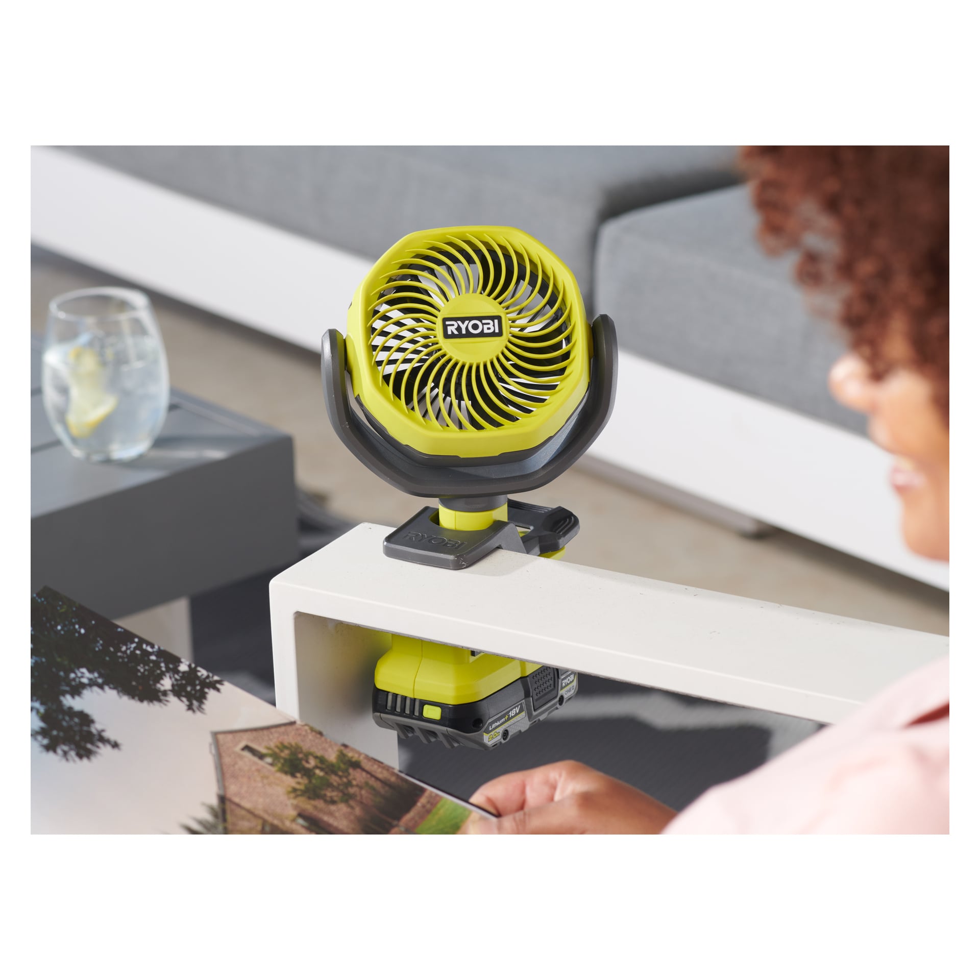 En person läser nära en liten, ljusgrön Ryobi One+ batteridriven bordsfläkt, fastklämd vid ett vitt bord. I bakgrunden finns ett glas isvatten och en grå soffa.