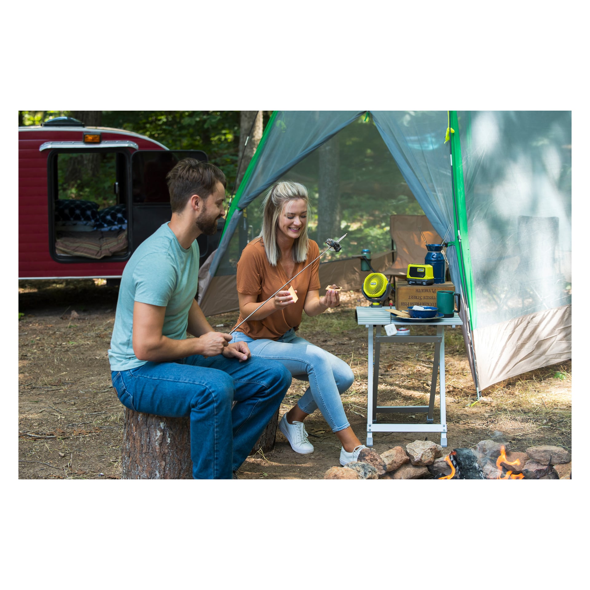 En man och en kvinna sitter vid en lägereld och rostar marshmallows bredvid sitt tält. I närheten finns ett litet bord med campingutrustning, muggar, Ryobi One+ batteridriven bordsfläkt RCF18-0 och en radio. I bakgrunden finns en röd husvagn omgiven av träd.