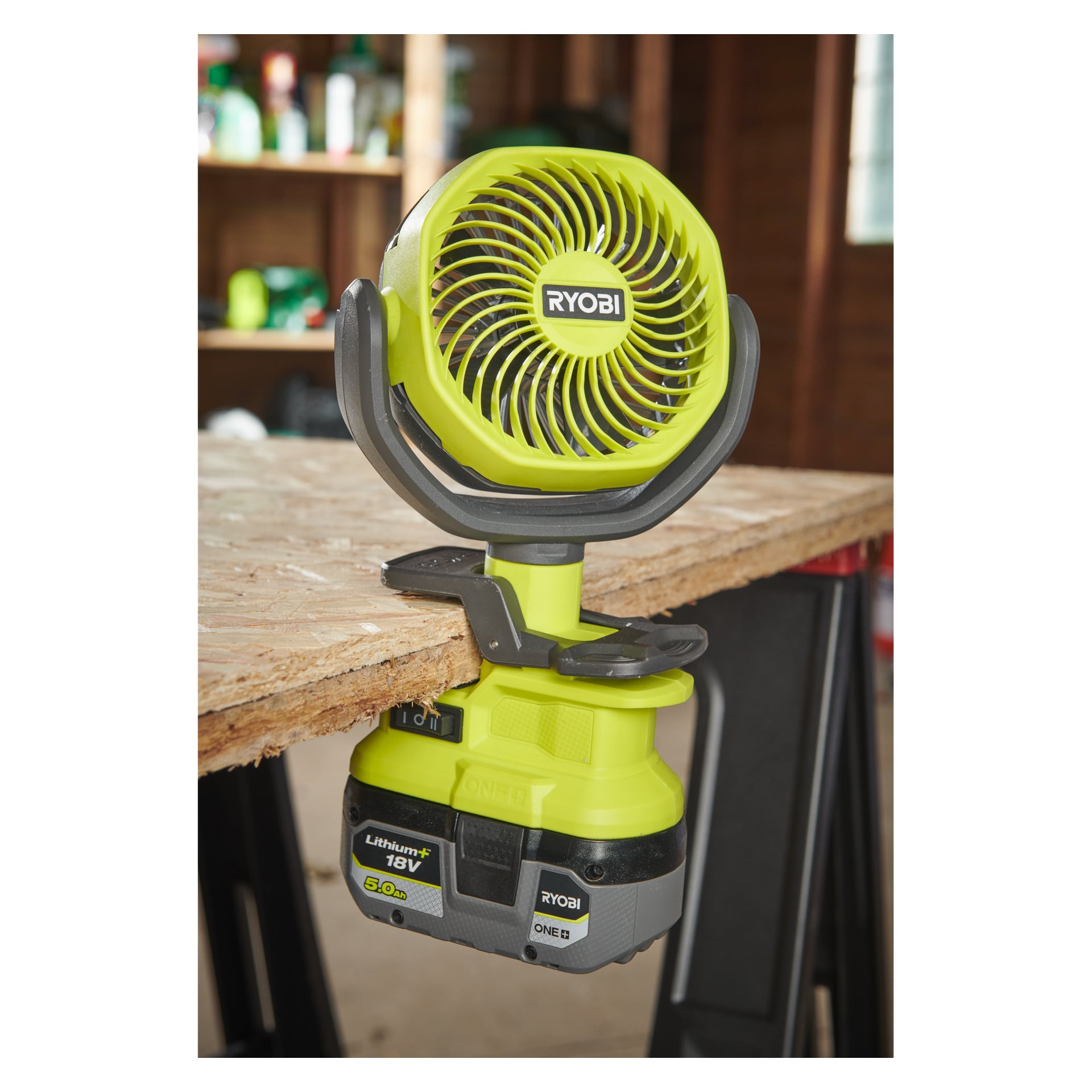 En Ryobi One+ RCF18-0 bordsfläkt, driven av ett 18V litiumbatteri, är fäst på sidan av en träskiva i en verkstad. Hyllor och olika verktyg syns i bakgrunden.