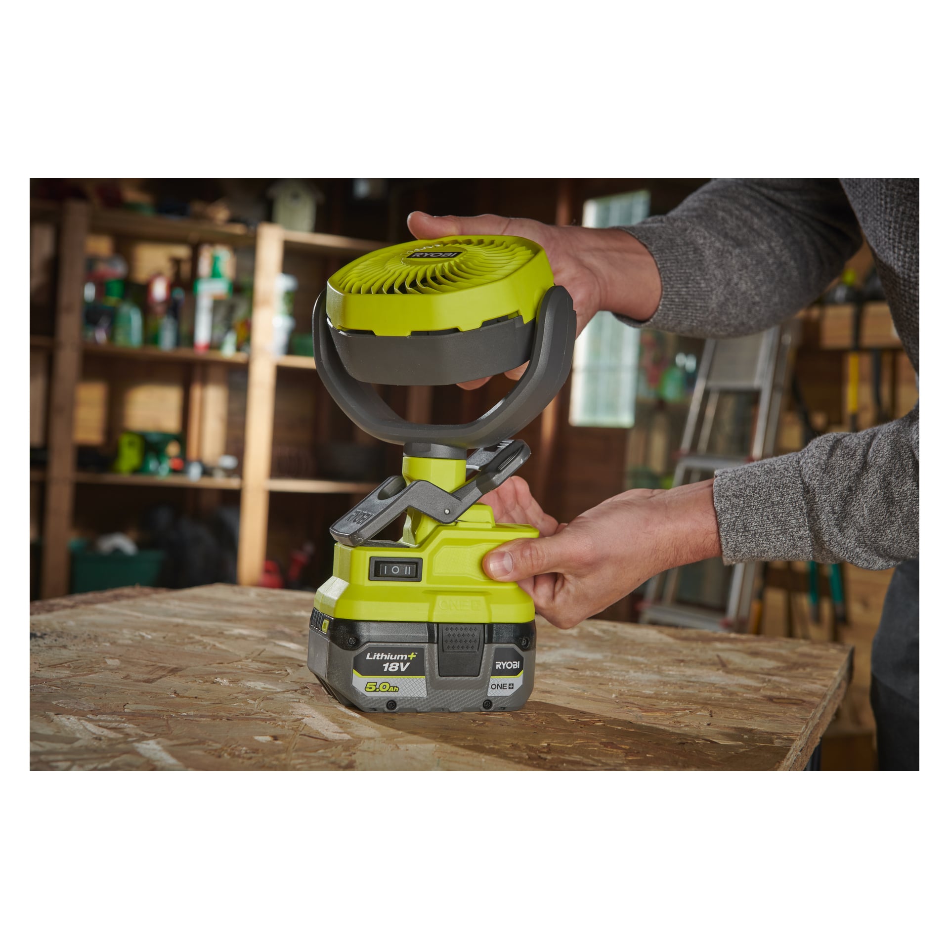 Någon justerar en Ryobi One+ RCF18-0 batteridriven bordsfläkt med ett 18V litiumjonbatteri, placerad på en träarbetsbänk i en verkstad. Hyllor fyllda med verktyg och utrustning syns bakom dem.