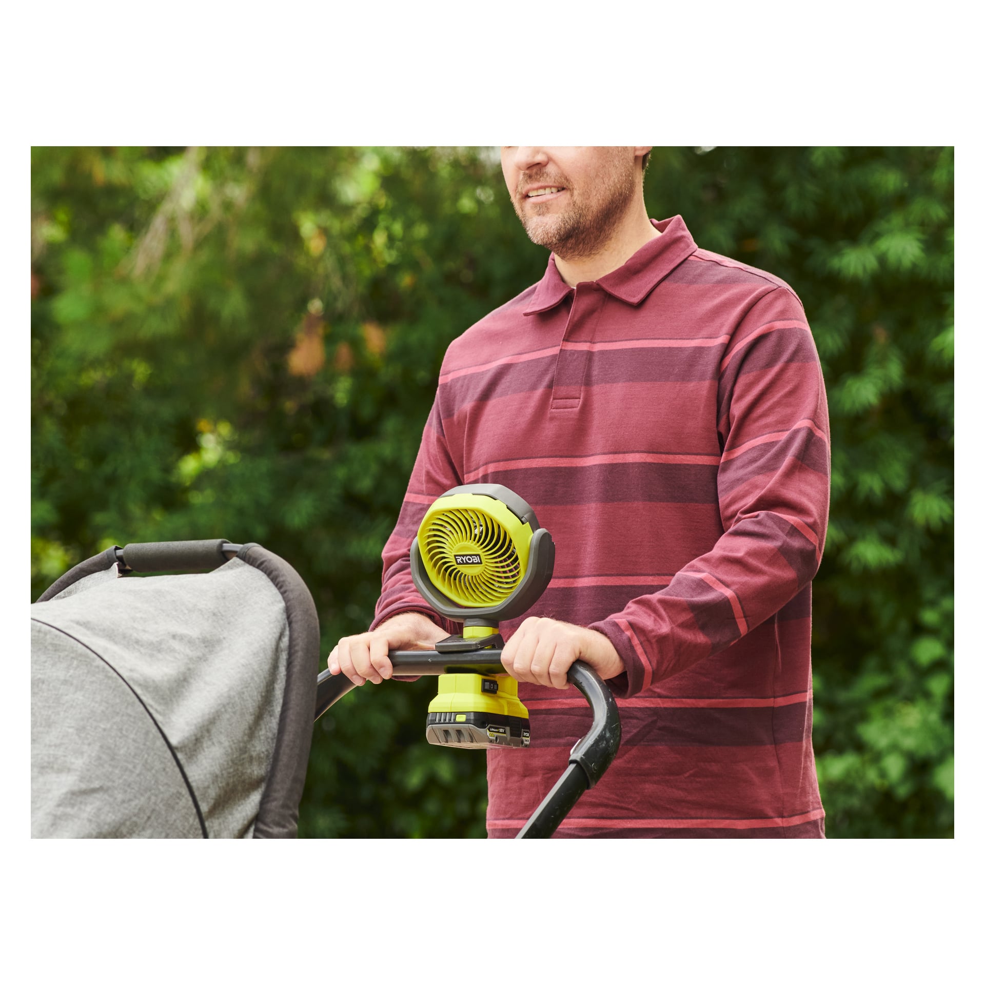En person med rödrandig tröja skjuter en barnvagn med en grön och svart Ryobi One+ batteridriven bordsfläkt (modell RCF18-0) monterad på styret. Frodigt grönt växtlighet kan ses i bakgrunden.