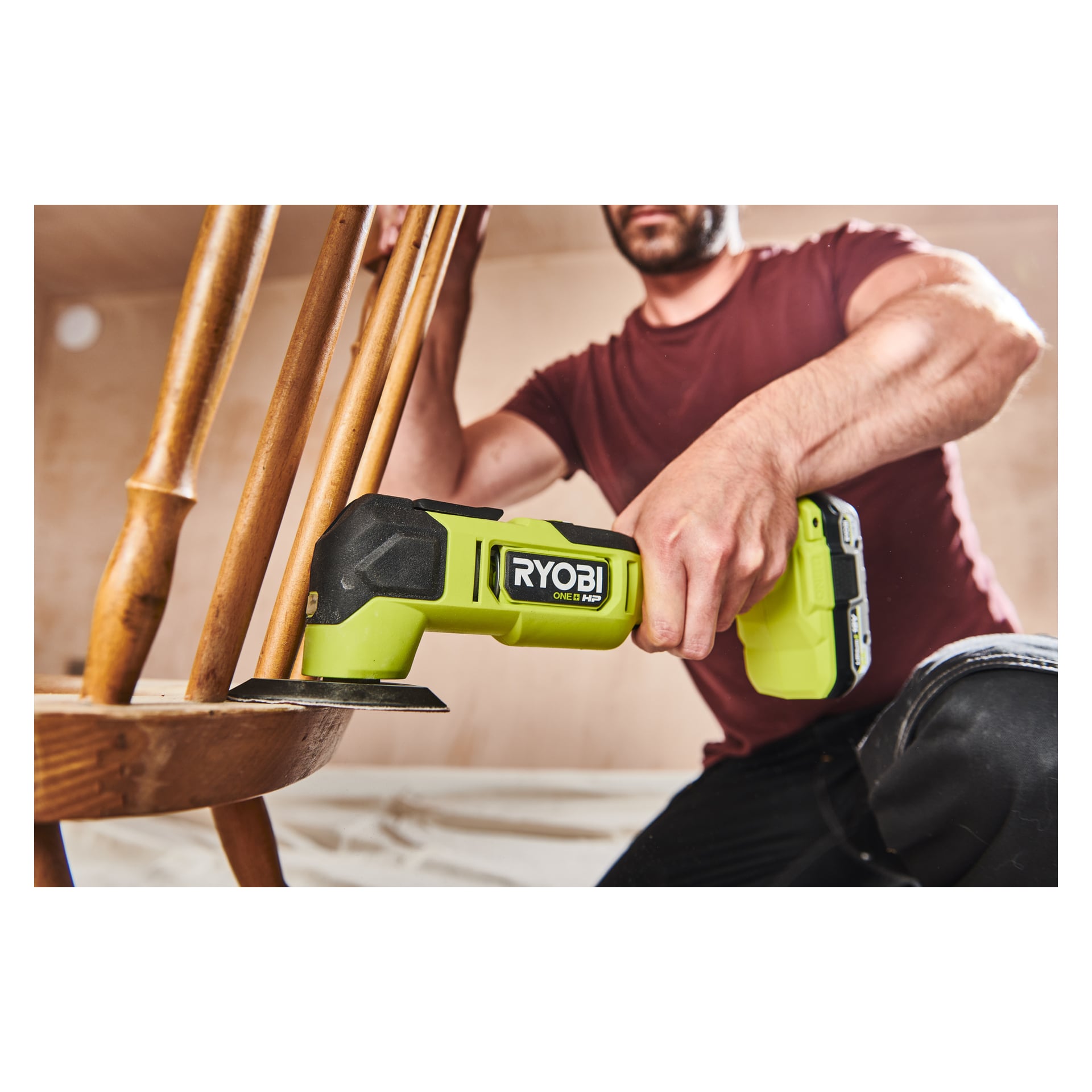 En person i en vinröd kortärmad skjorta knäböjer inomhus och arbetar på träbenet på en stol med en grön Ryobi One+ HP Multi-tool RMT18X-0.