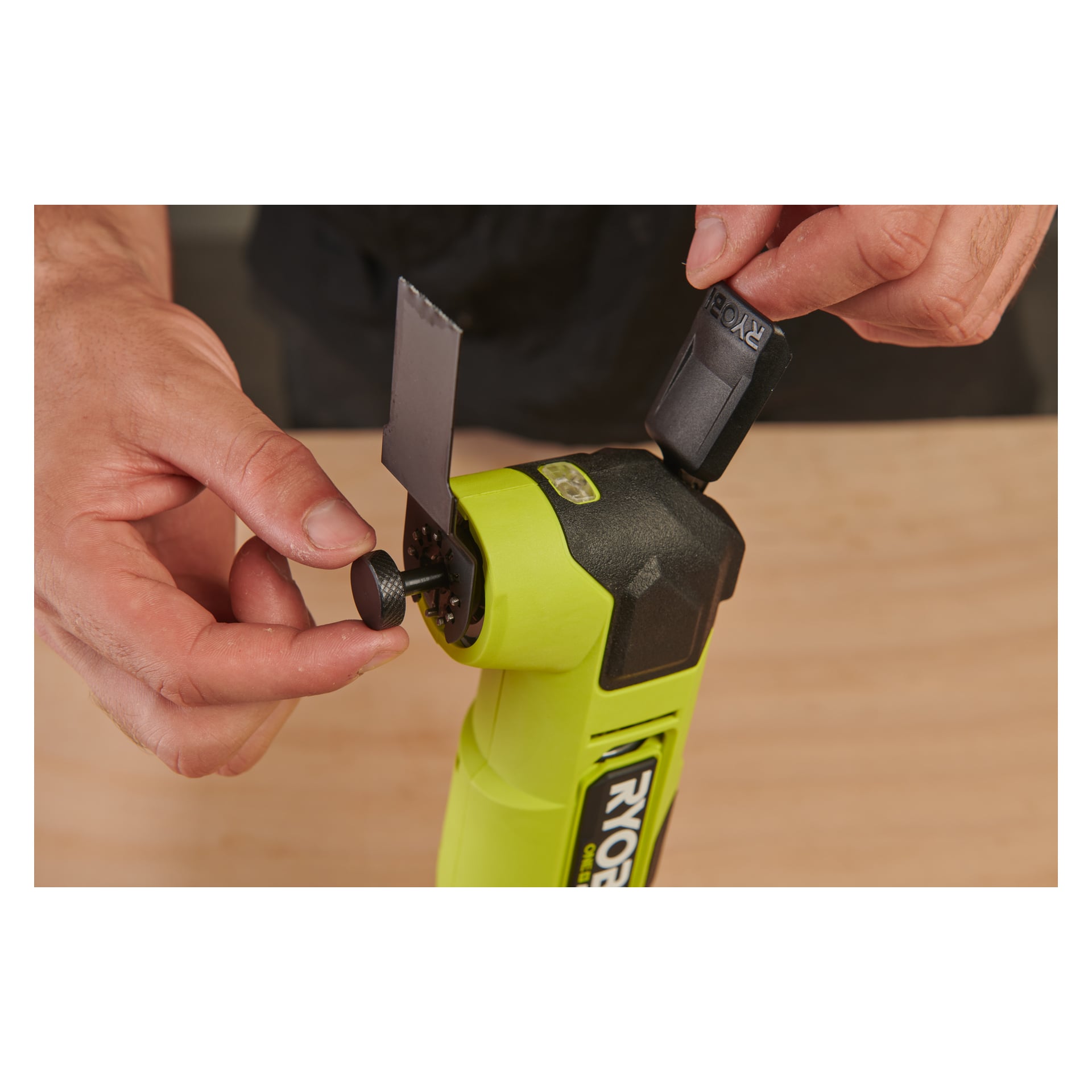 Någon fäster ett metallblad på en Ryobi One+ HP Multitool RMT18X-0 med en svart skruv, och håller verktyget stadigt på en blek träyta.