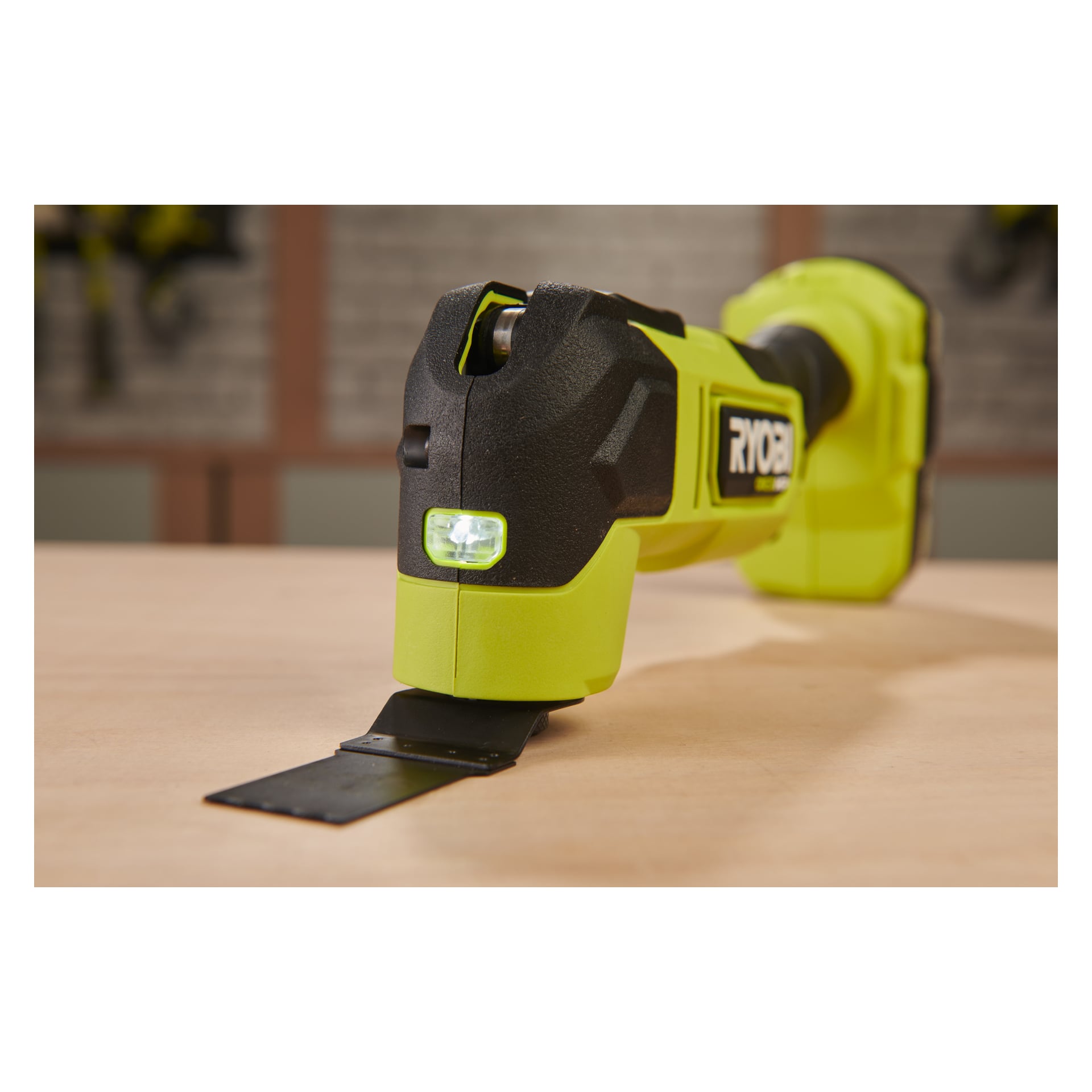 En grön och svart Ryobi One+ HP RMT18X-0 sladdlös oscillerande multitool visas på nära håll, liggande på en träyta med sitt platta metallblad pekande nedåt.