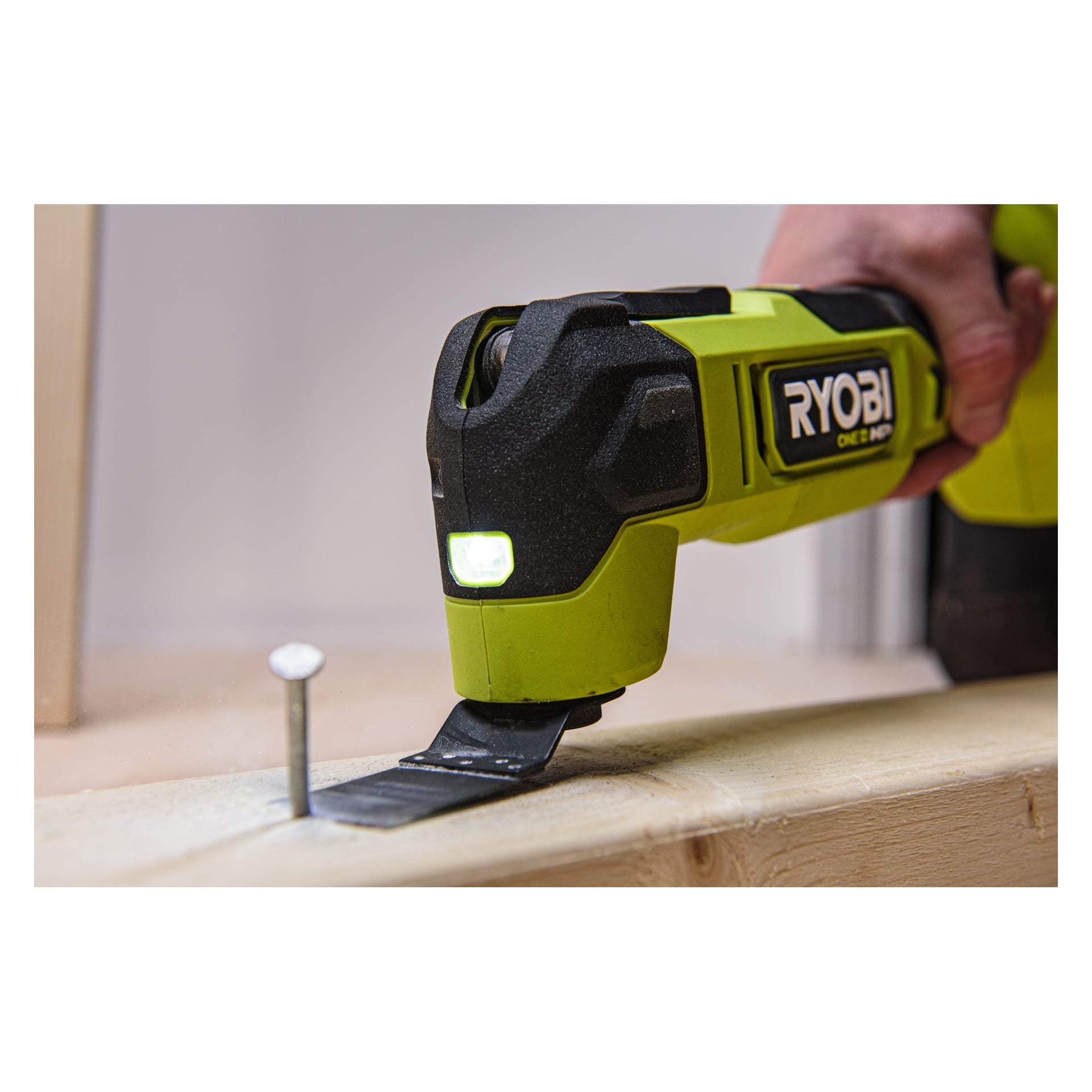 En hand håller en Ryobi One+ HP Multitool RMT18X-0 utrustad med ett platt blad, som skär i en träskiva nära en upprätt spik.