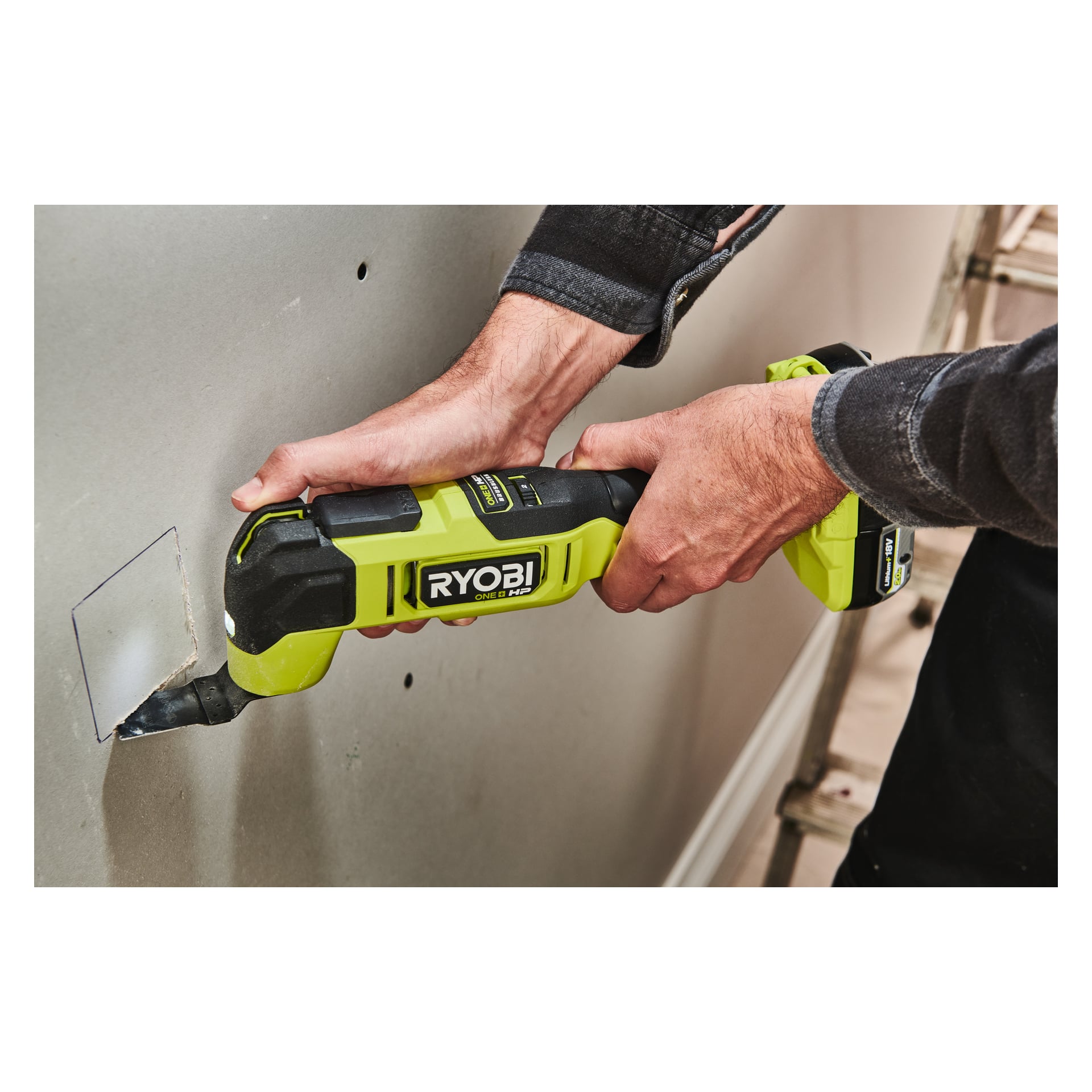 Någon använder en grön Ryobi One+ HP Multitool RMT18X-0 med båda händerna för att skära ut en rektangulär öppning i gipsskivan, eftersom sektionen som ska skäras är delvis losstagen.