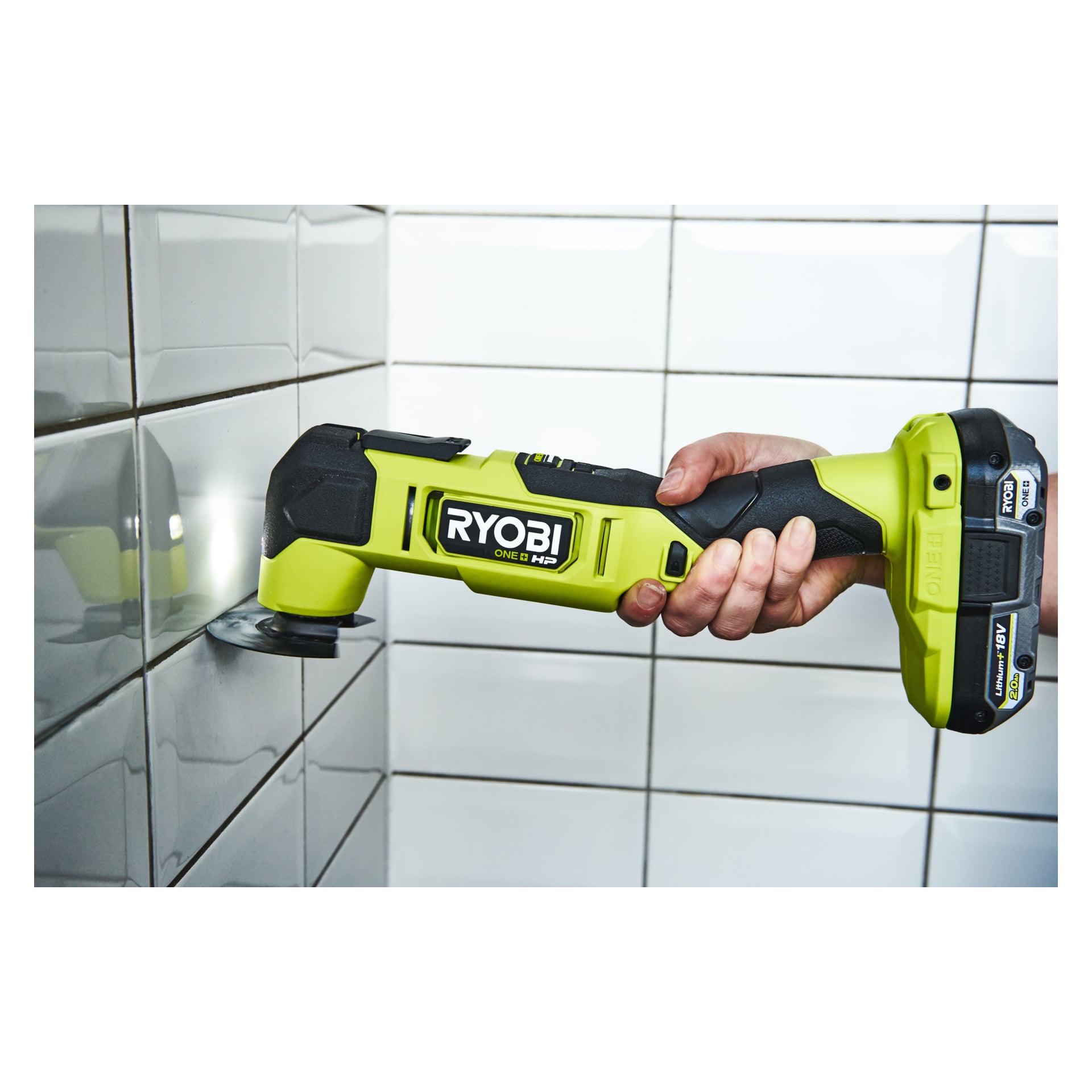 En hand håller i en grön Ryobi One+ HP Multitool RMT18X-0 utrustad med ett skärblad, hållet mot kaklade väggar och verkar användas för att skära eller ta bort fogar mellan plattorna.