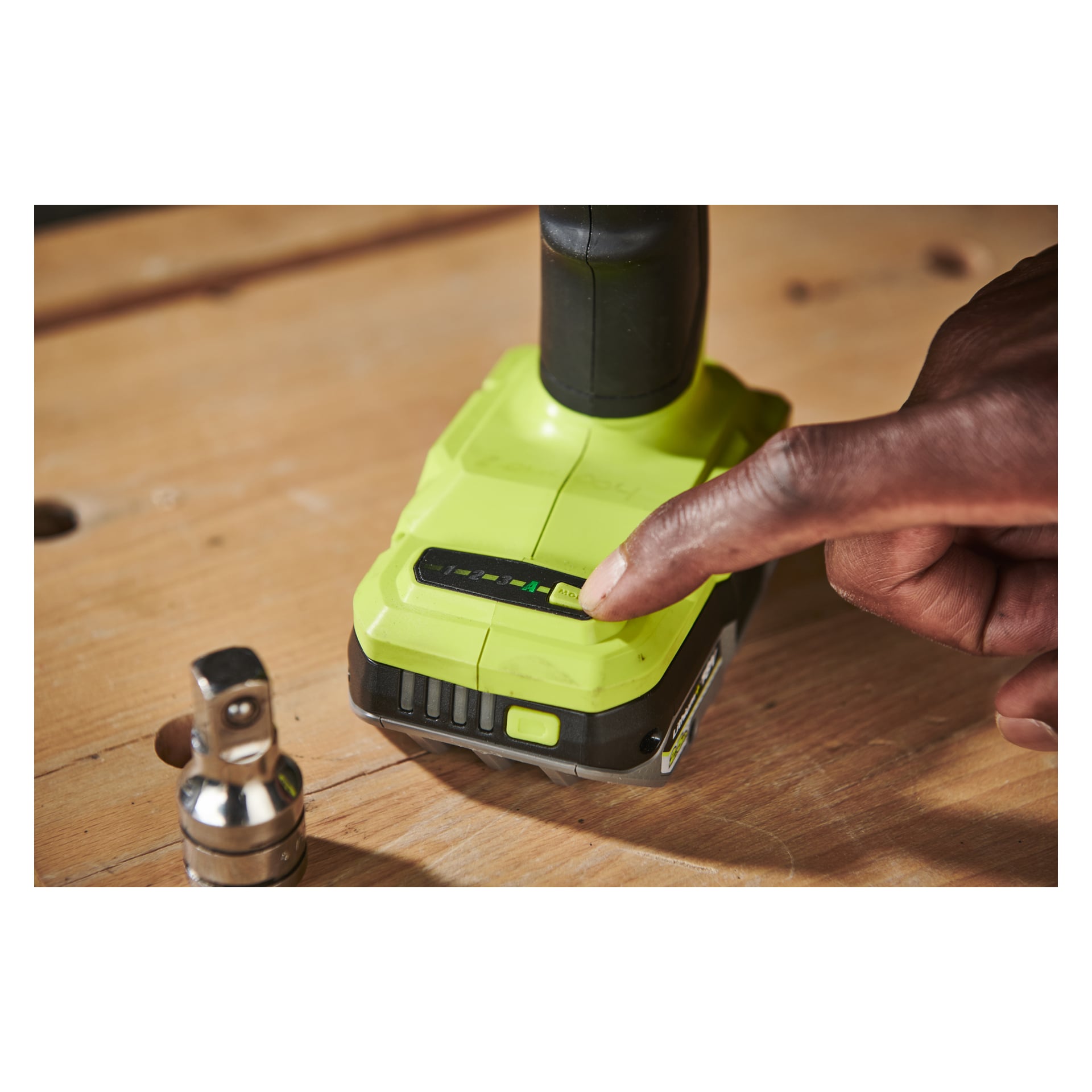 Sormi painaa akun ilmaisinpainiketta vihreässä Ryobi One+ HP Impact Wrench RIW18C-0 -paristossa, joka on asetettu puupinnalle, ja lähellä on pieni metallinen pistorasia.