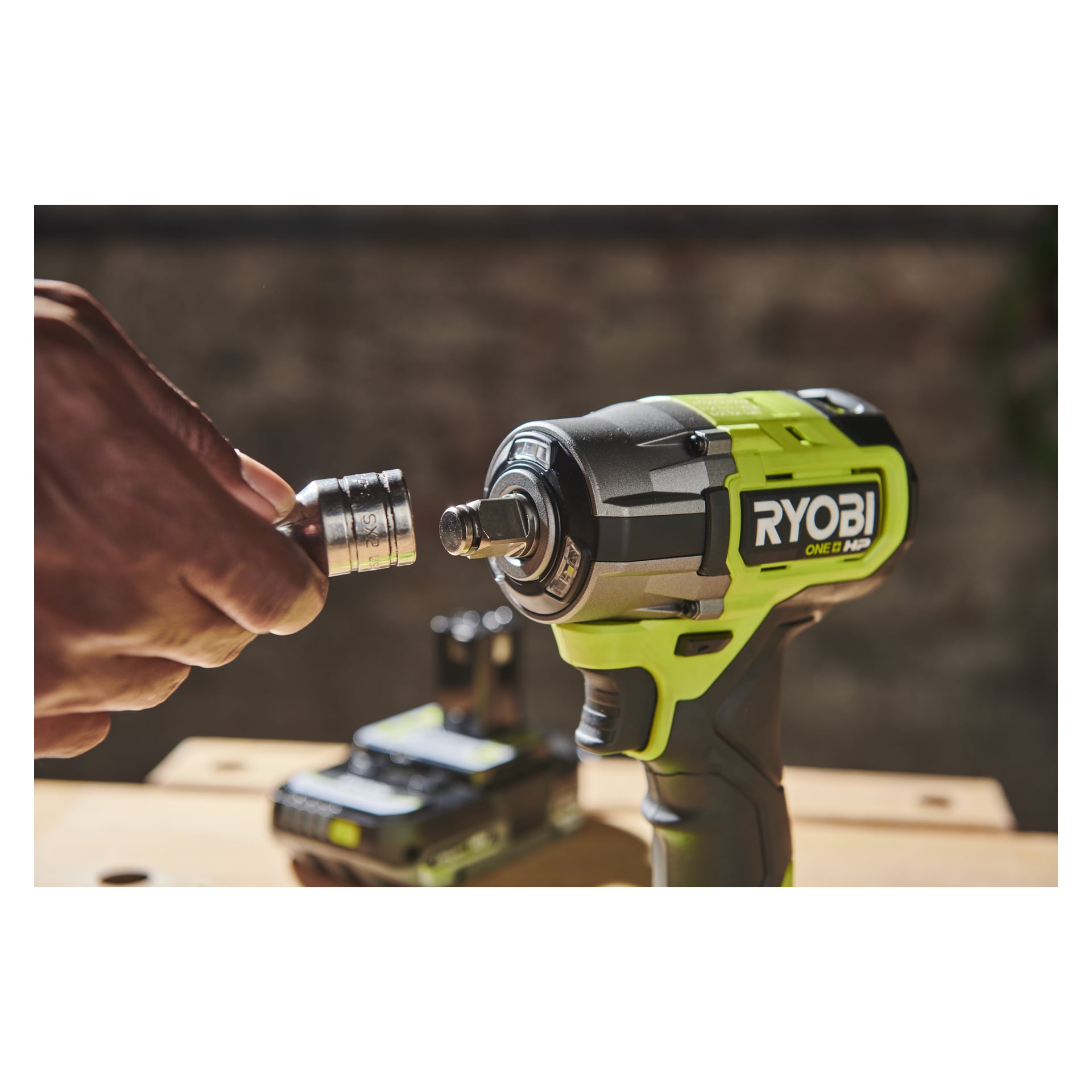 Joku asentaa hylsyn Ryobi One+ HP Impact Wrench RIW18C-0:aan; Taustalla työpöydällä näkyy akku ja laturi epätarkennuksena.