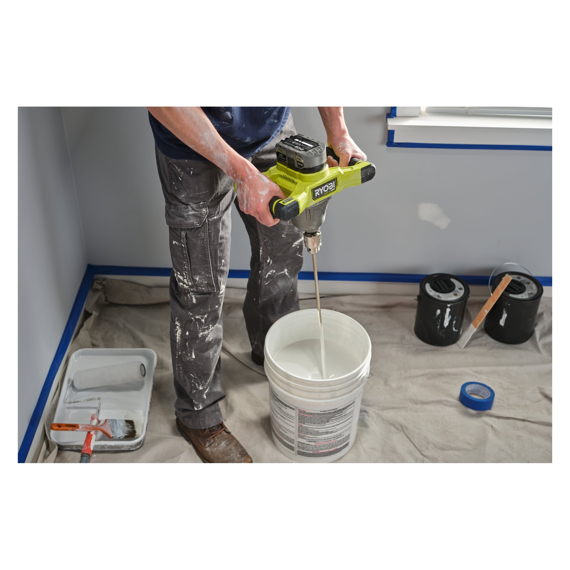 En person blandar färg i en vit hink med en Ryobi One+ HP Agitator RPM18X-0. Golvet skyddas av en duk, med färgburkar, en rullbricka och tejp placerade i närheten.