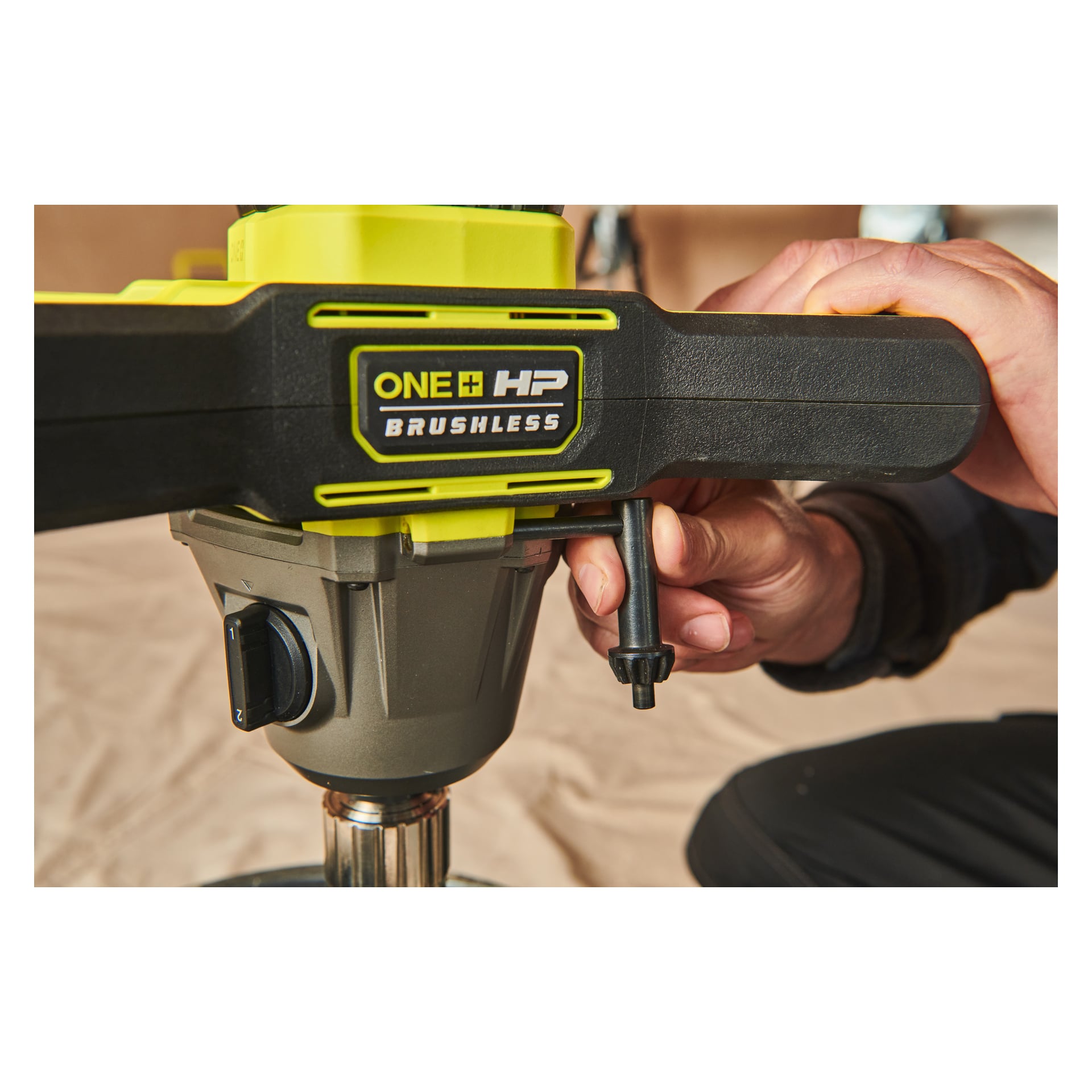En närbild visar en hand som vrider på en ratt på ett Ryobi One+ HP Agitator RPM18X-0 elverktyg, med en beige tygyta i närheten och bakgrunden mjukt ur fokus.