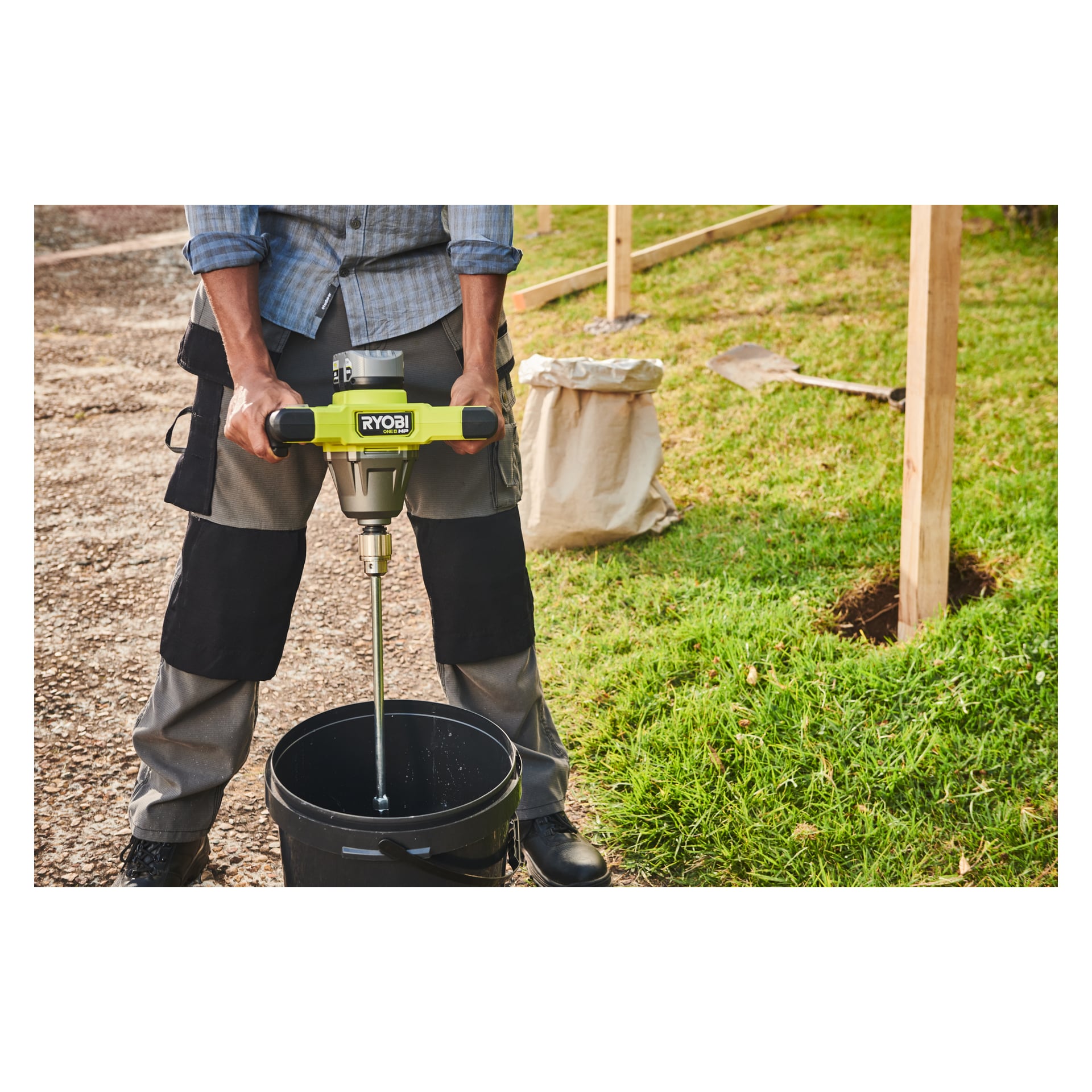 En person blandar material i en svart hink med en Ryobi One+ HP Agitator RPM18X-0 utomhus på grus. I närheten finns en spade, en papperspåse och några trästolpar bredvid gräset.
