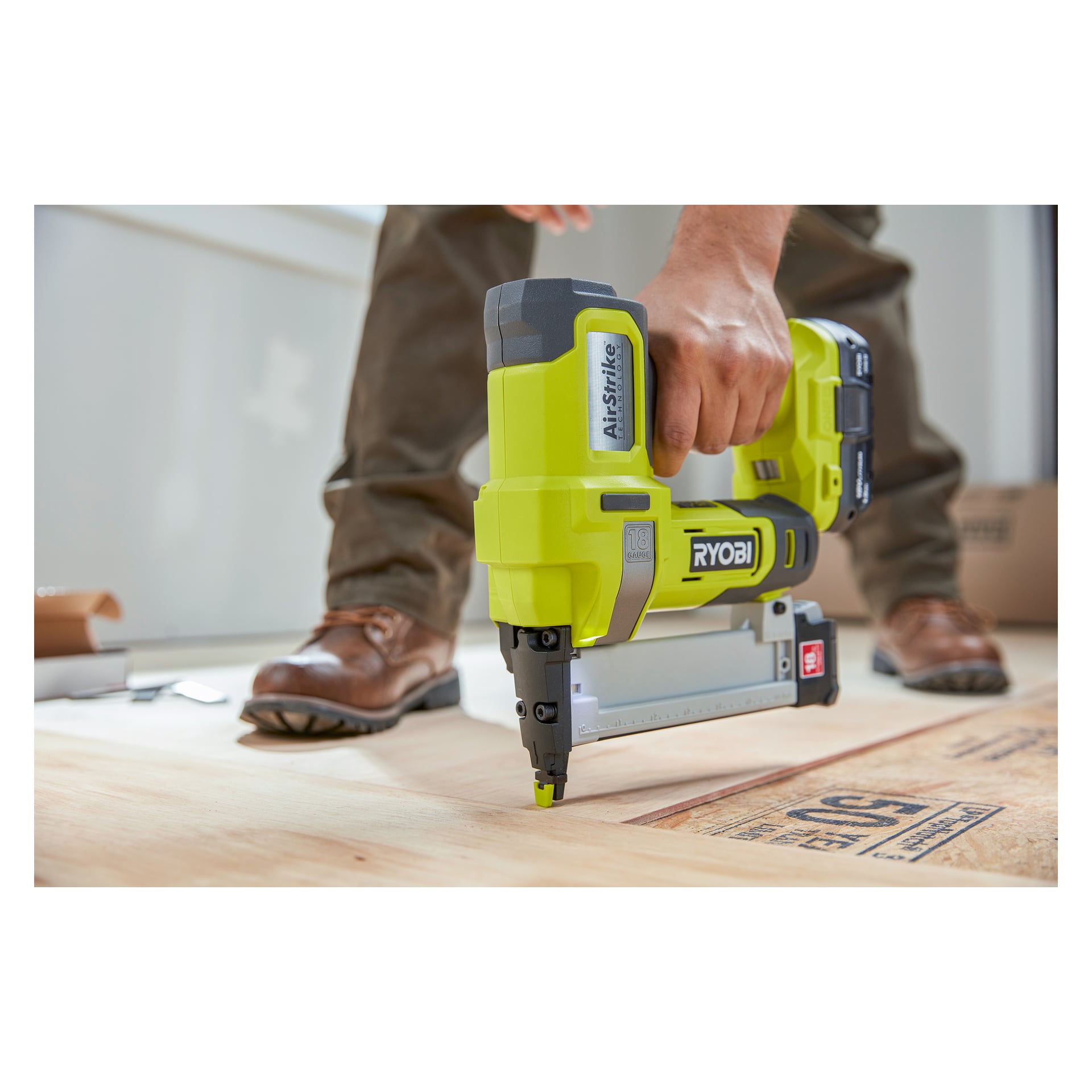 En hand manövrerar en Ryobi One+ Staple Gun R18GS18-0 för att fästa trägolvet, med hand, verktyg och stövlar visade. Fokus ligger på häftpistolen när den trycks mot golvet.