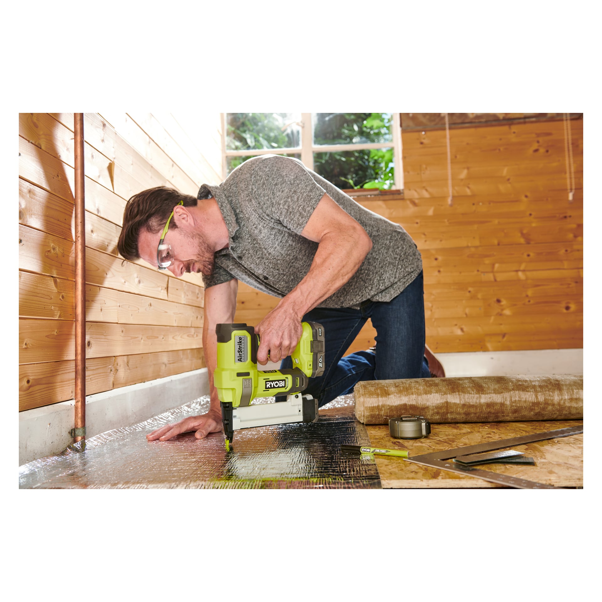 En man knäböjer på golvet i ett trärum och fäster reflekterande isolering med en Ryobi One+ Staple Gun R18GS18-0. Olika verktyg och byggmaterial ligger utspridda i närheten.