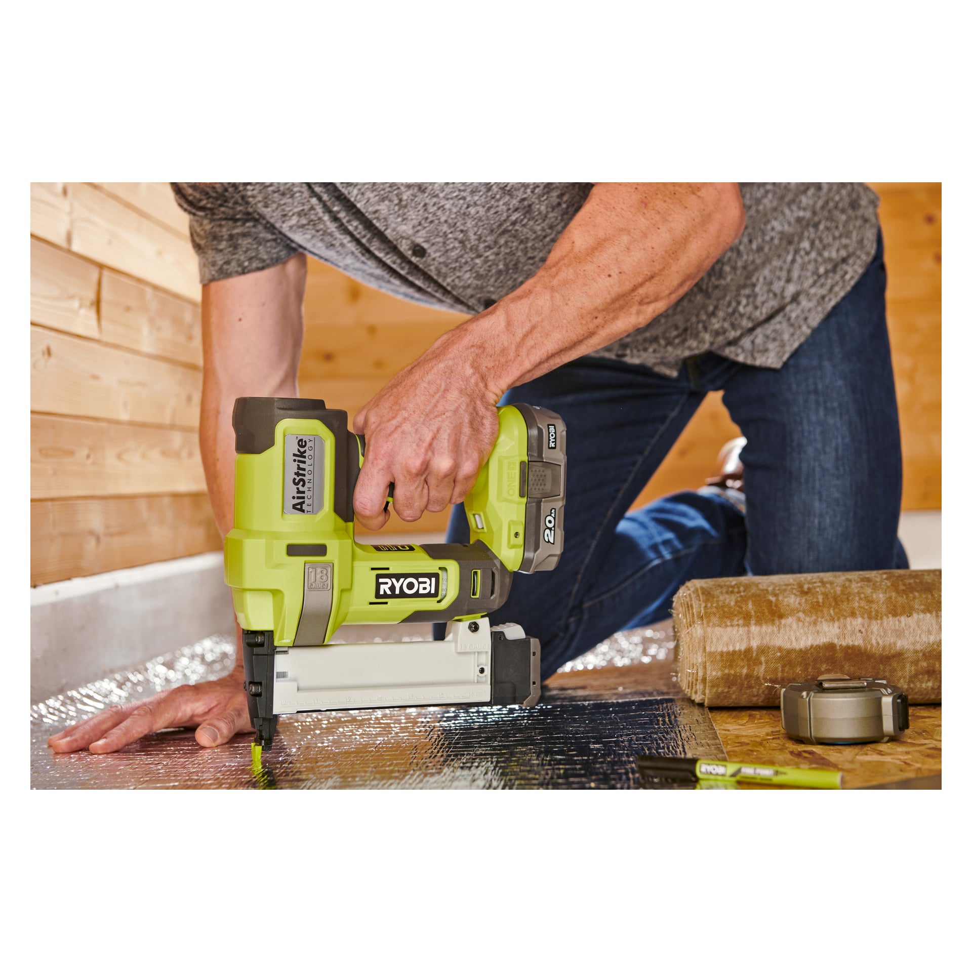 Någon använder en Ryobi One+ Staple Gun R18GS18-0 för att fästa material på ett golv med reflekterande isolering. Deras hand trycker ner på verktyget, med ett batteripaket och en rulle matta i närheten.