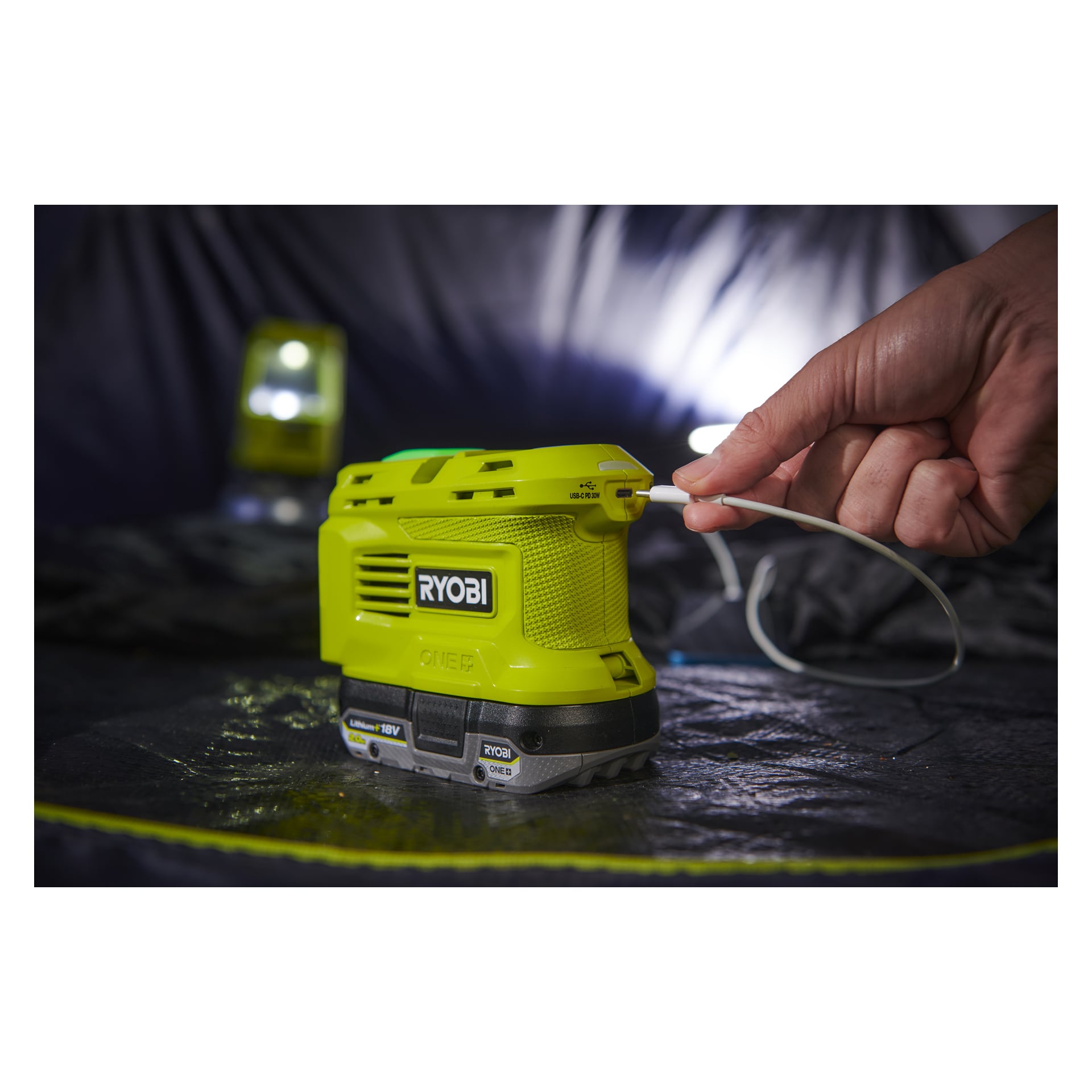 En hand kopplar in en kabel i en grön Ryobi One+ spänningsomvandlare märkt "RY18BI150B" på en mörk yta, med en glödande lykta mjukt suddig i bakgrunden.
