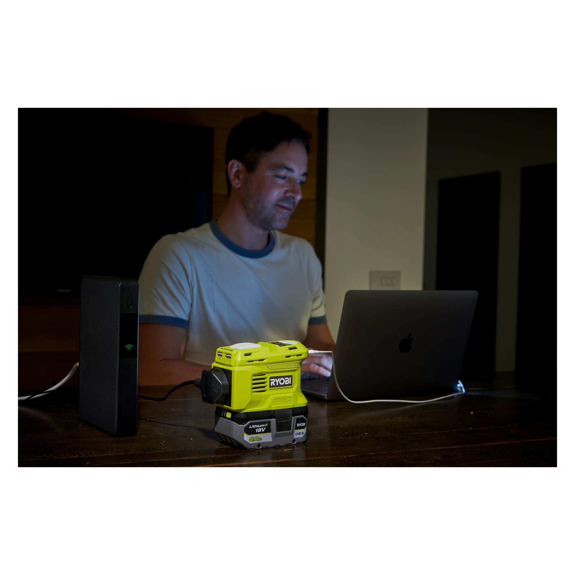 En man använder en laptop vid ett bord, driven av en portabel powerbank och spänningsomvandlaren RY18BI150B från Ryobi One+, båda med indikatorlampor tända.