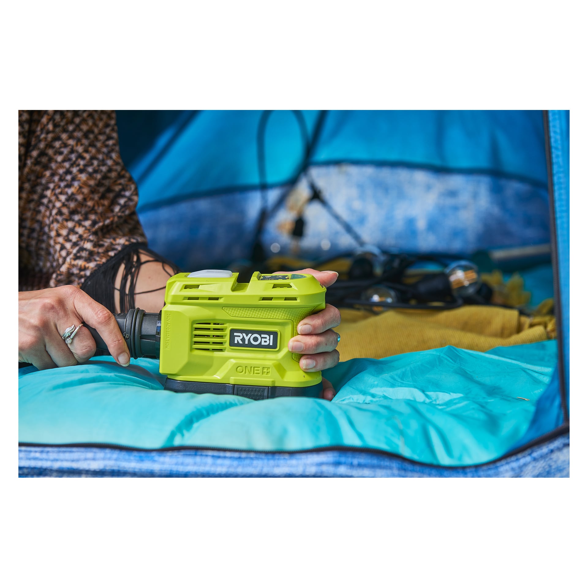 En hand styr en grön "RY18BI150B"-spänningsomvandlare från Ryobi One+ för att blåsa upp en blå luftmadrass inne i ett tält dekorerat med ljusslingor och en gul filt.