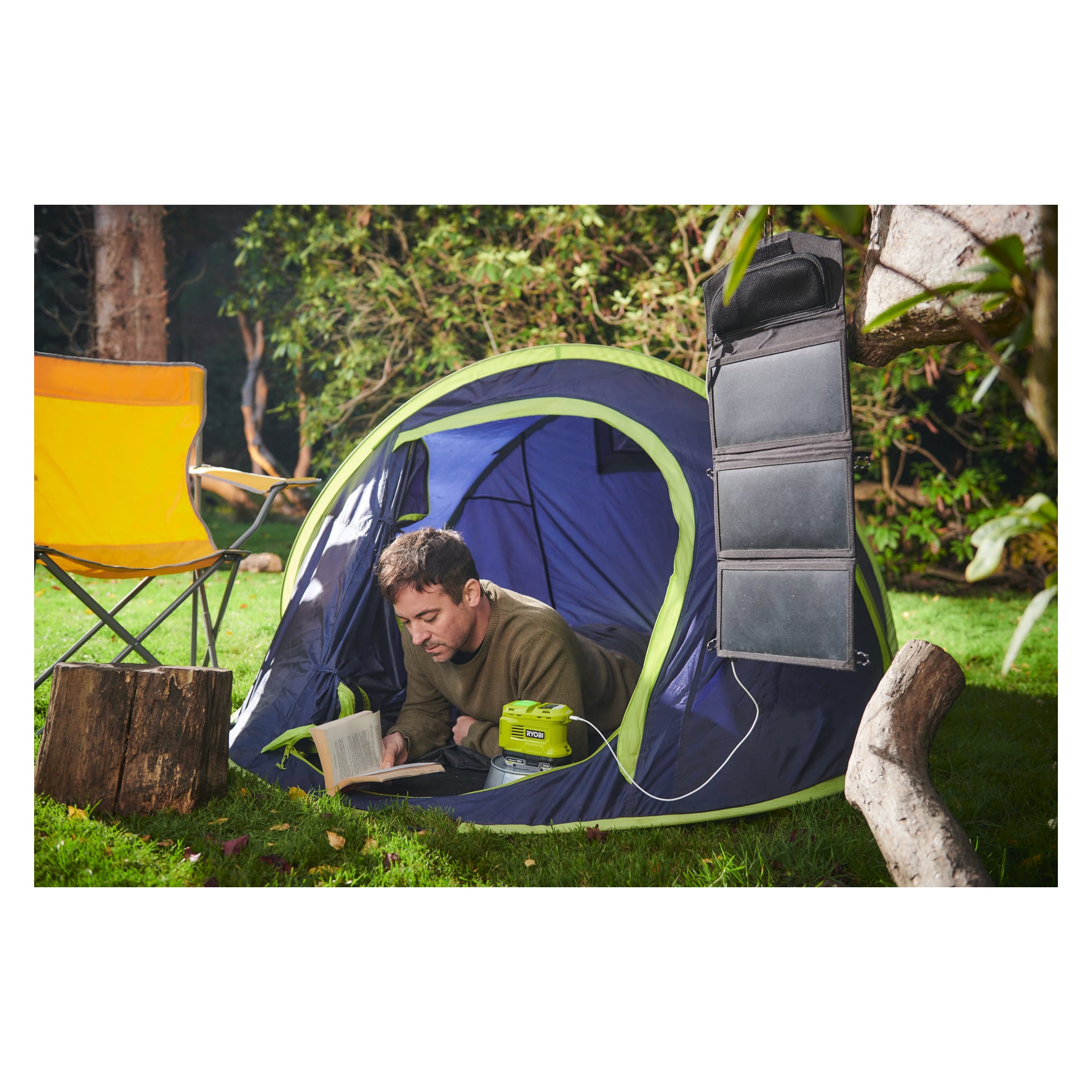 En man slappnar av i ett blått och grönt tält och läser en bok. Bredvid tältet på den gräsbevuxna campingplatsen står en gul stol, en solpanel och en Ryobi One+ spänningsomvandlare märkt "RY18BI150B" i bruk.