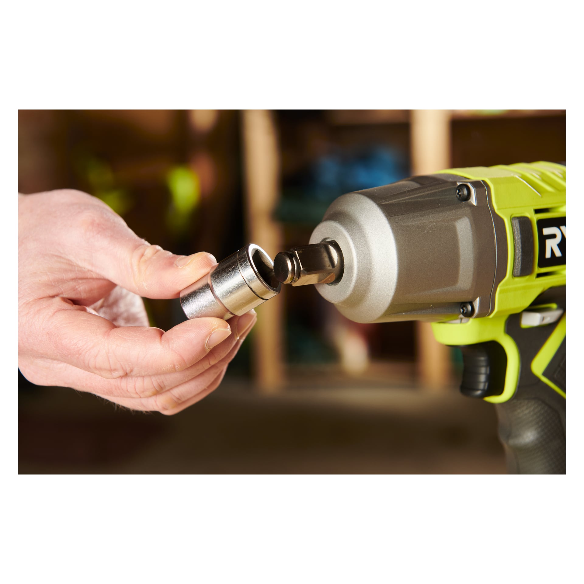 Någon monterar en metallhylsa på den fyrkantiga disken på en Ryobi One+ Impact Wrench RIW18-0, igenkännbar på sitt gröna och svarta färgschema, inne i en verkstad.