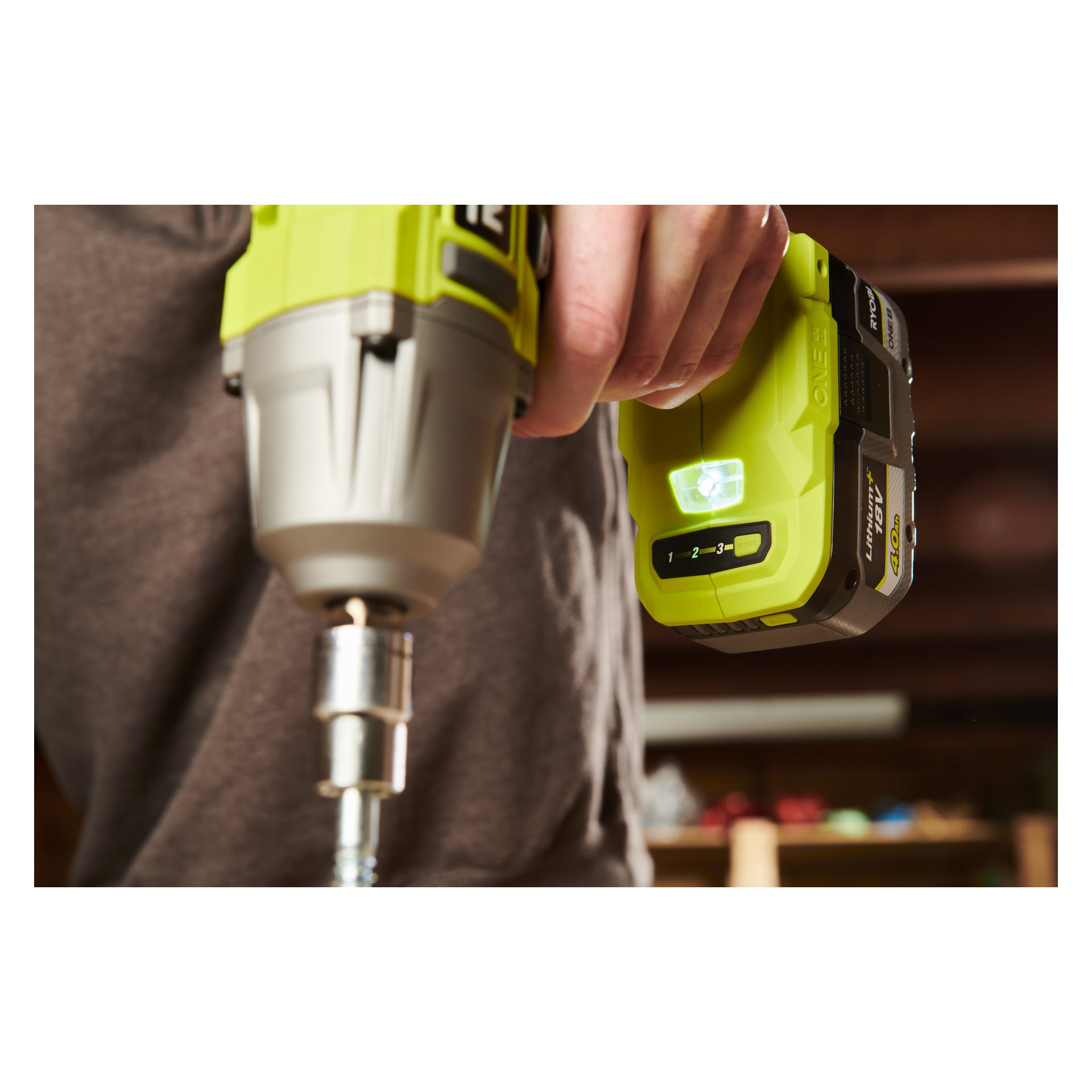 Någon håller i en grön Ryobi One+ slagnyckel RIW18-0, fingret trycker på batteriindikatorknappen. Verktyget svävar ovanför en polerad metallskruv, med en mjukt oskarp bakgrund.