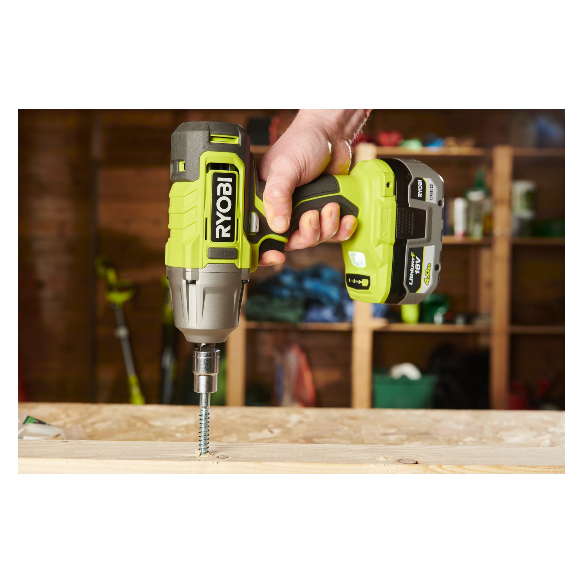 Någon använder en grön och svart Ryobi One+ Impact Wrench RIW18-0 för att fästa en stor skruv i en träskiva, med hyllor fyllda med verktyg och material synliga i verkstadens bakgrund.
