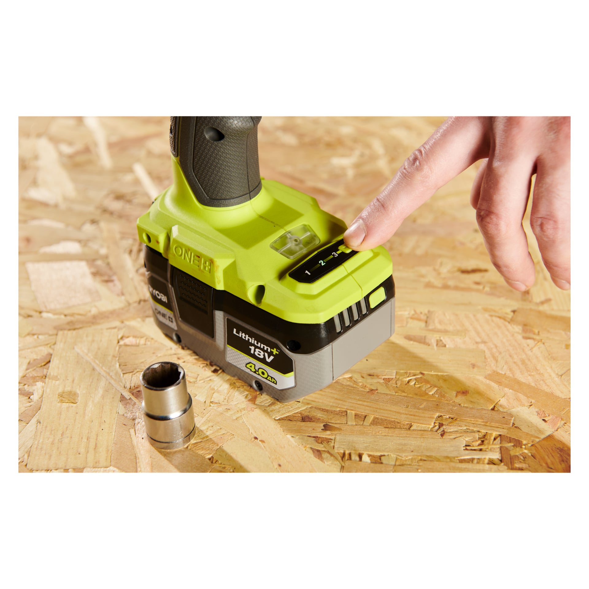 En hand trycker på batterinivåindikatorn på ett grönt Ryobi One+ 18V litiumbatteri kopplat till en Impact Wrench RIW18-0, vilket visar den aktuella laddningen. Bredvid den vilar en metallhylsa på en träyta.