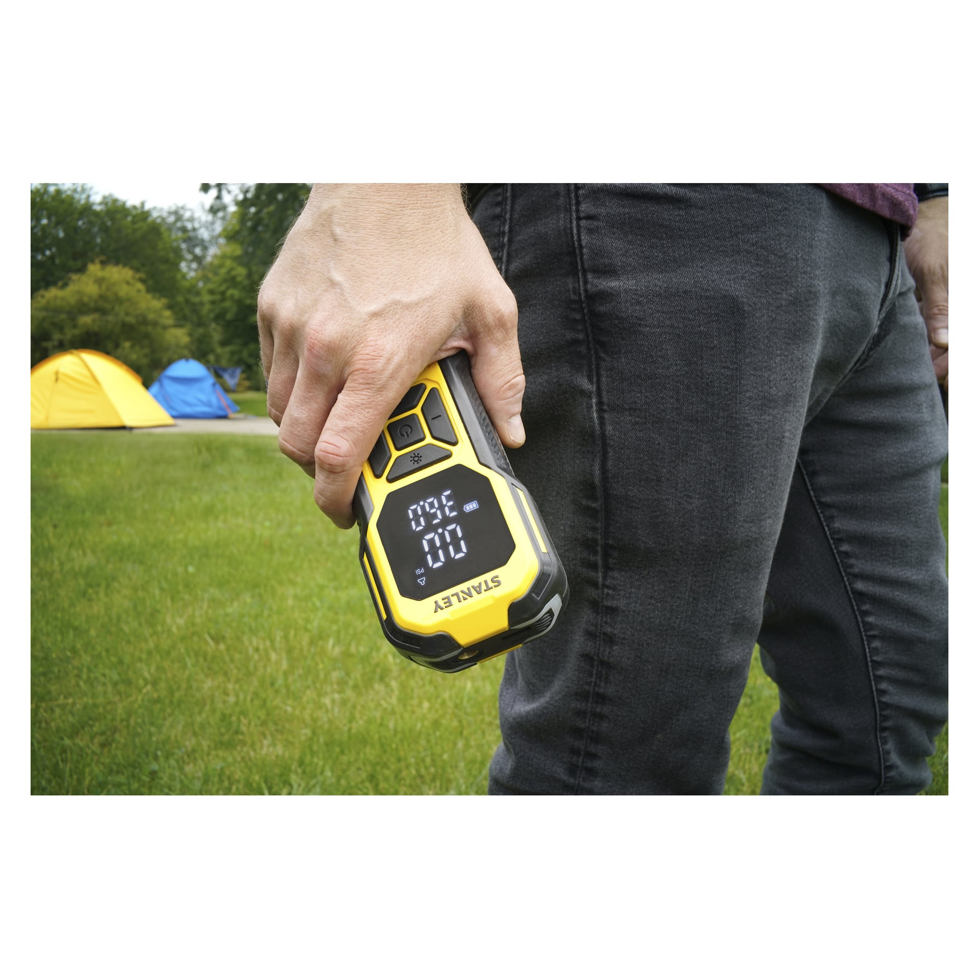 Osoba ubrana w ciemne dżinsy i bordową koszulkę stoi na trawie, trzymając SXIF0101 Stanley Rechargeable Mini Air Pump z widocznym cyfrowym wyświetlaczem. Za nimi znajdują się dwa namioty i kilka drzew.