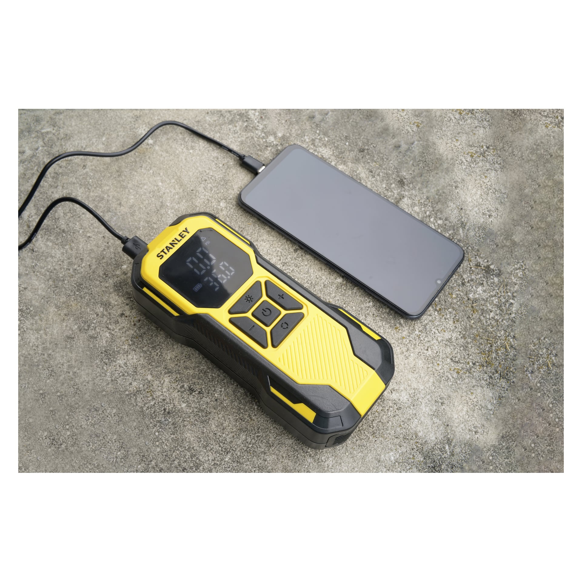Żółto-miniaturowa pompa powietrza Stanley Rechargeable SXIF0101 z cyfrowym wyświetlaczem jest podłączona do smartfona na teksturowanej, szarej powierzchni. Na ekranie wyświetlane są różne liczby i ikony.