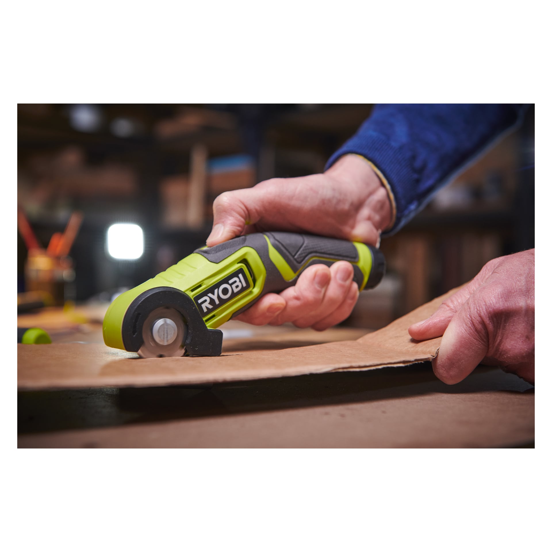 Någon klipper en bit kartong med en grön och svart Ryobi USB Lithium multi-scissor RCT4-0 i en verkstad eller i garage.