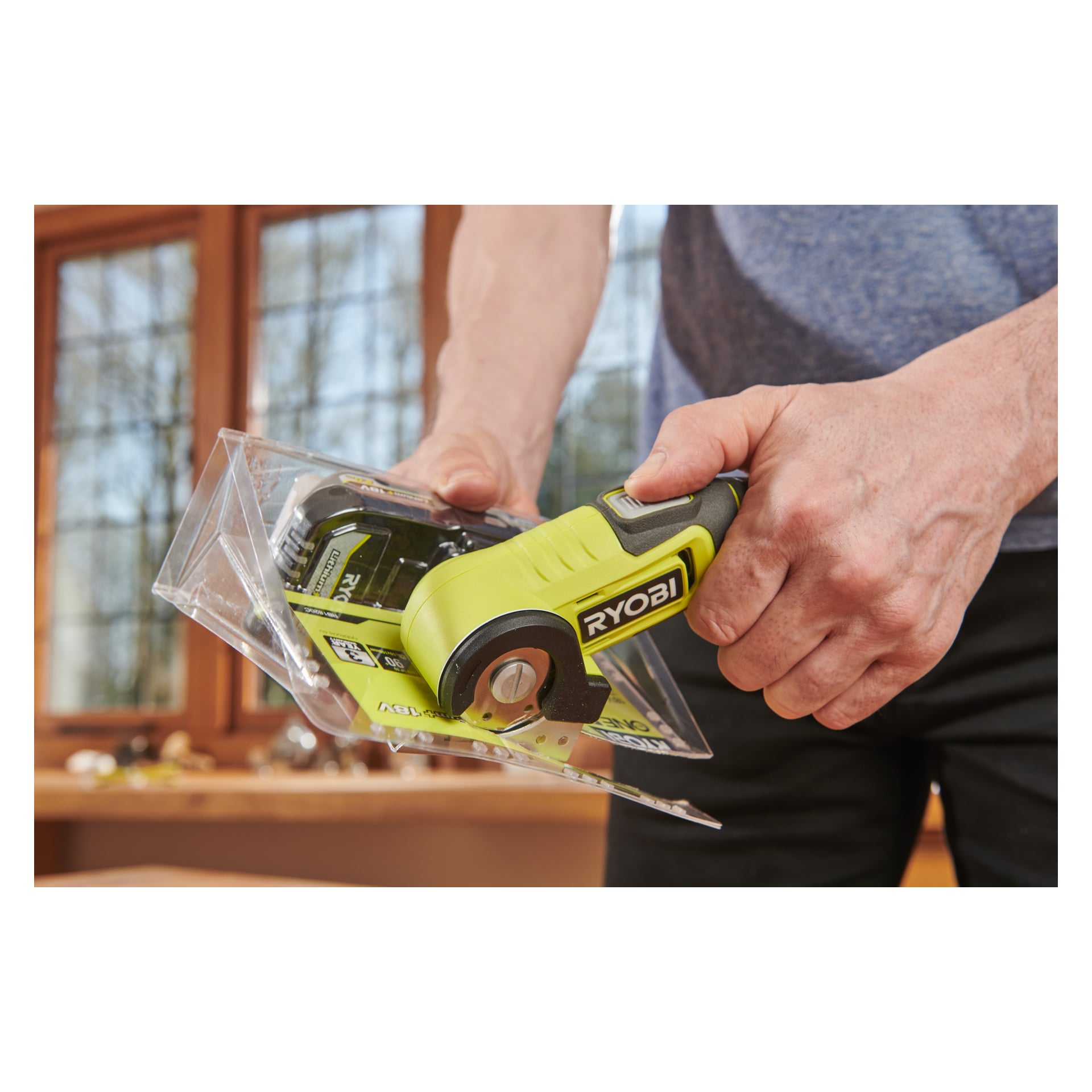 Någon öppnar plastförpackningar inomhus med en Ryobi USB Lithium Battery-driven multi-sax RCT4-0, och stora fönster kan ses i bakgrunden.