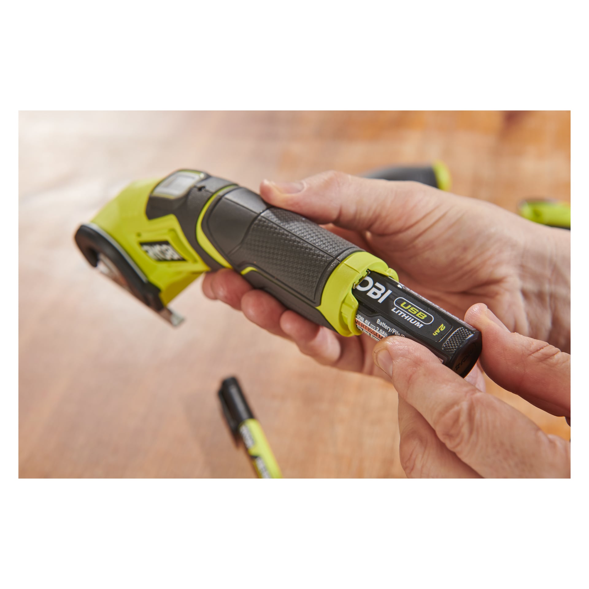 Någon placerar ett Ryobi USB Lithium-uppladdningsbart batteri i en grön och svart RCT4-0 batteridriven multisax, med ett träbord synligt bakom.