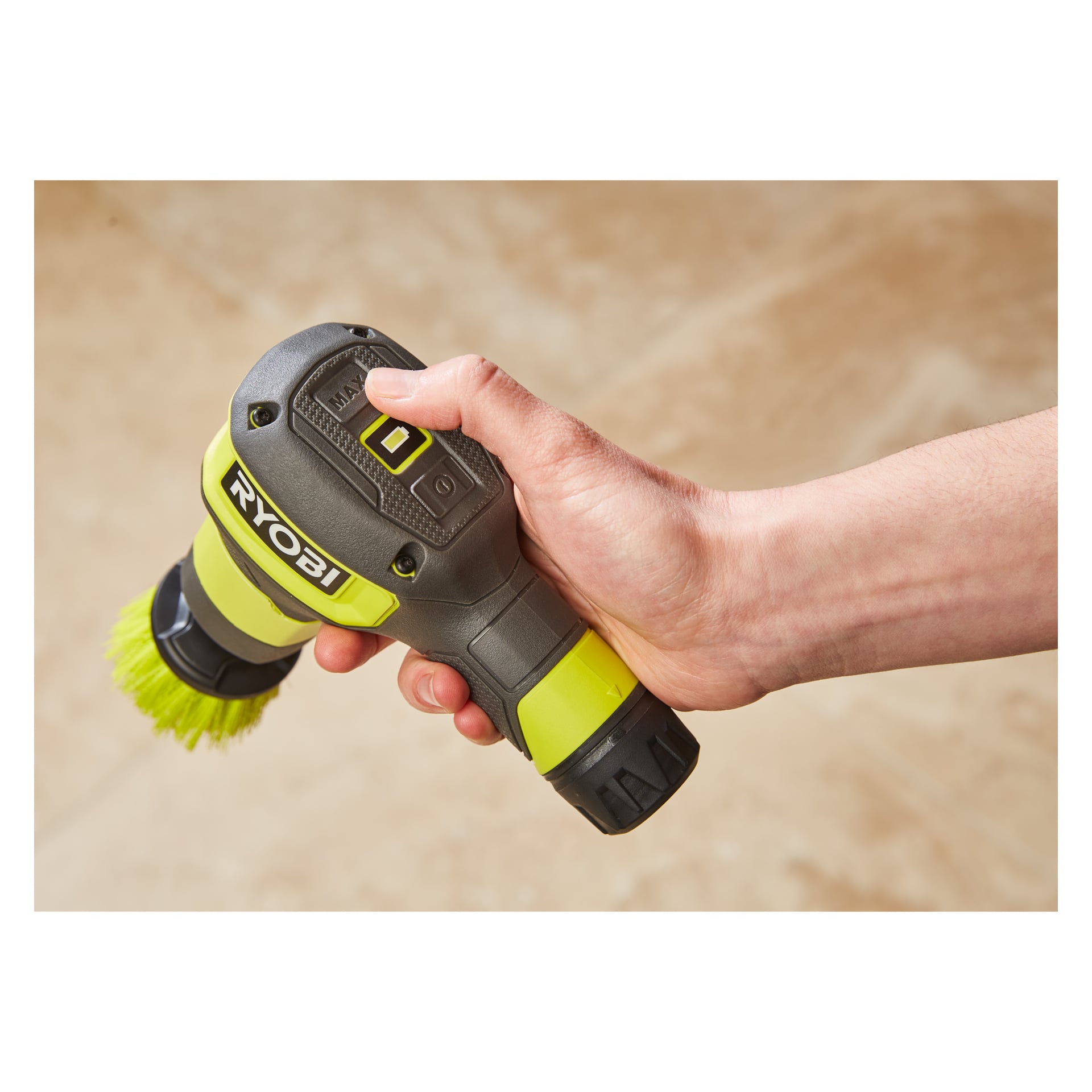 Eine Person hält eine Ryobi USB Lithium Rotierende Waschbürste RSCR4-0 mit einer angehängten grünen Bürste, die den Power-Knopf und das sichtbare Ryobi-Logo auf einem hellbraunen Boden hervorhebt.