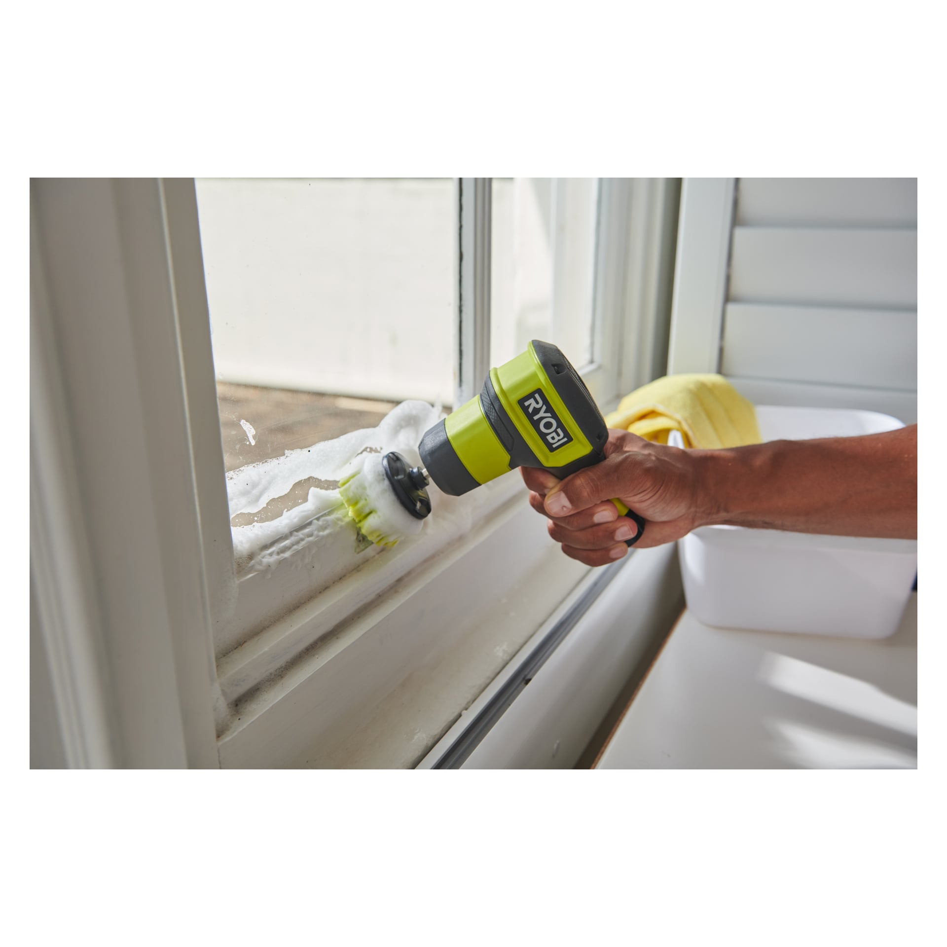 Jemand benutzt die Ryobi USB Lithium Rotating Wash Brush RSCR4-0, um einen Fensterrahmen zu schrubben, wodurch sichtbare Seifenschaum entsteht. Ein gelbes Tuch und ein weißer Behälter liegen nahe beieinander auf einer Oberfläche.
