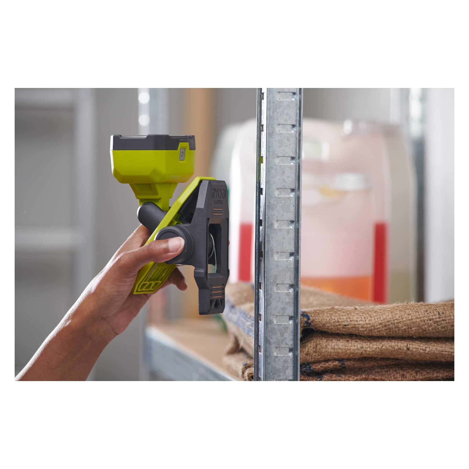 En person håller en grön och svart Ryobi USB Lithium Battery-driven arbetslampa (LED RLC4-0) framför en metallhylla. Bredvid hyllorna är säckar staplade bredvid en stor plastbehållare med orange vätska.