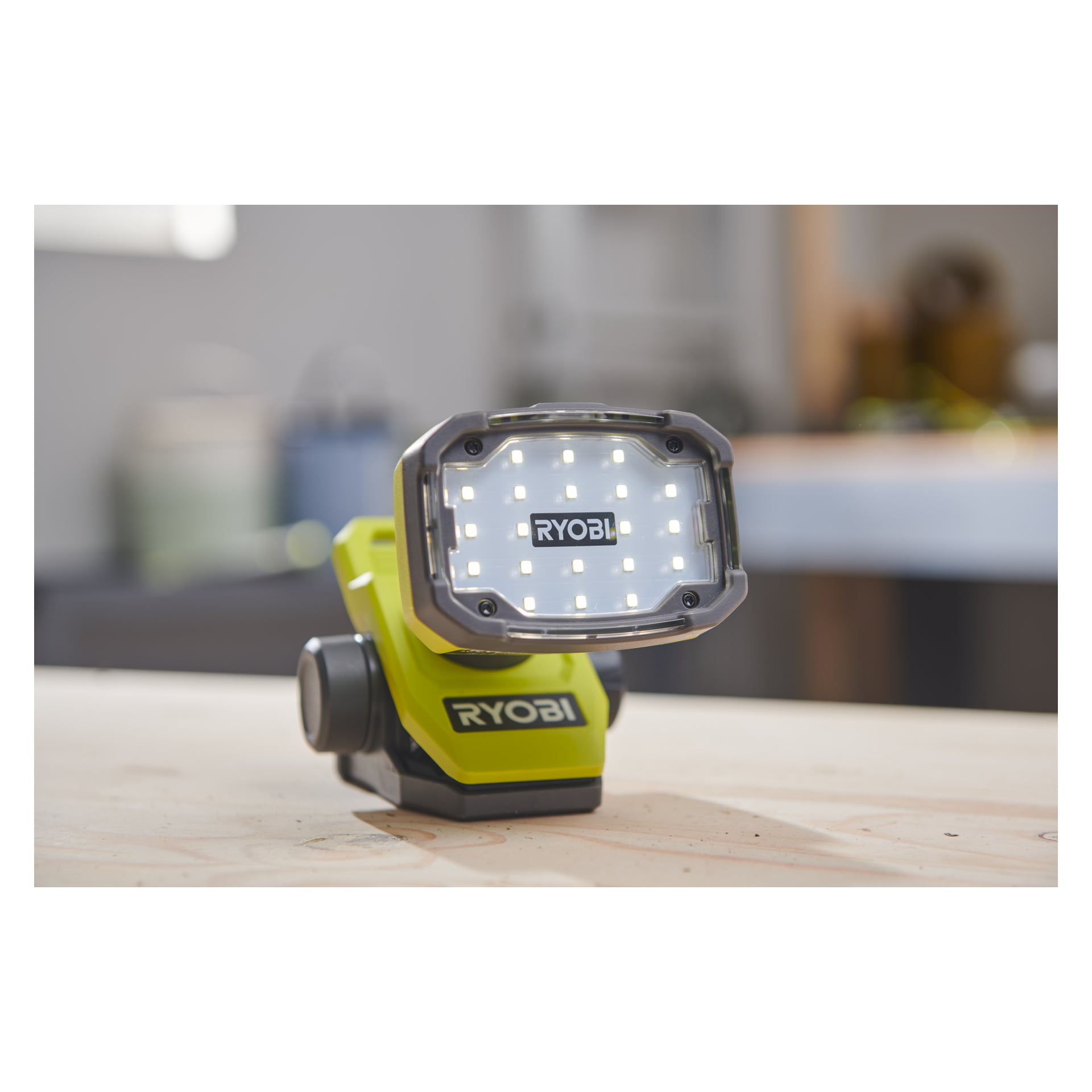 En grön och svart Ryobi USB Lithium Battery arbetslampa, modell RLC4-0, med flera lysande LED-lampor. Lampan står på en träyta i en verkstad, med synliga märken.