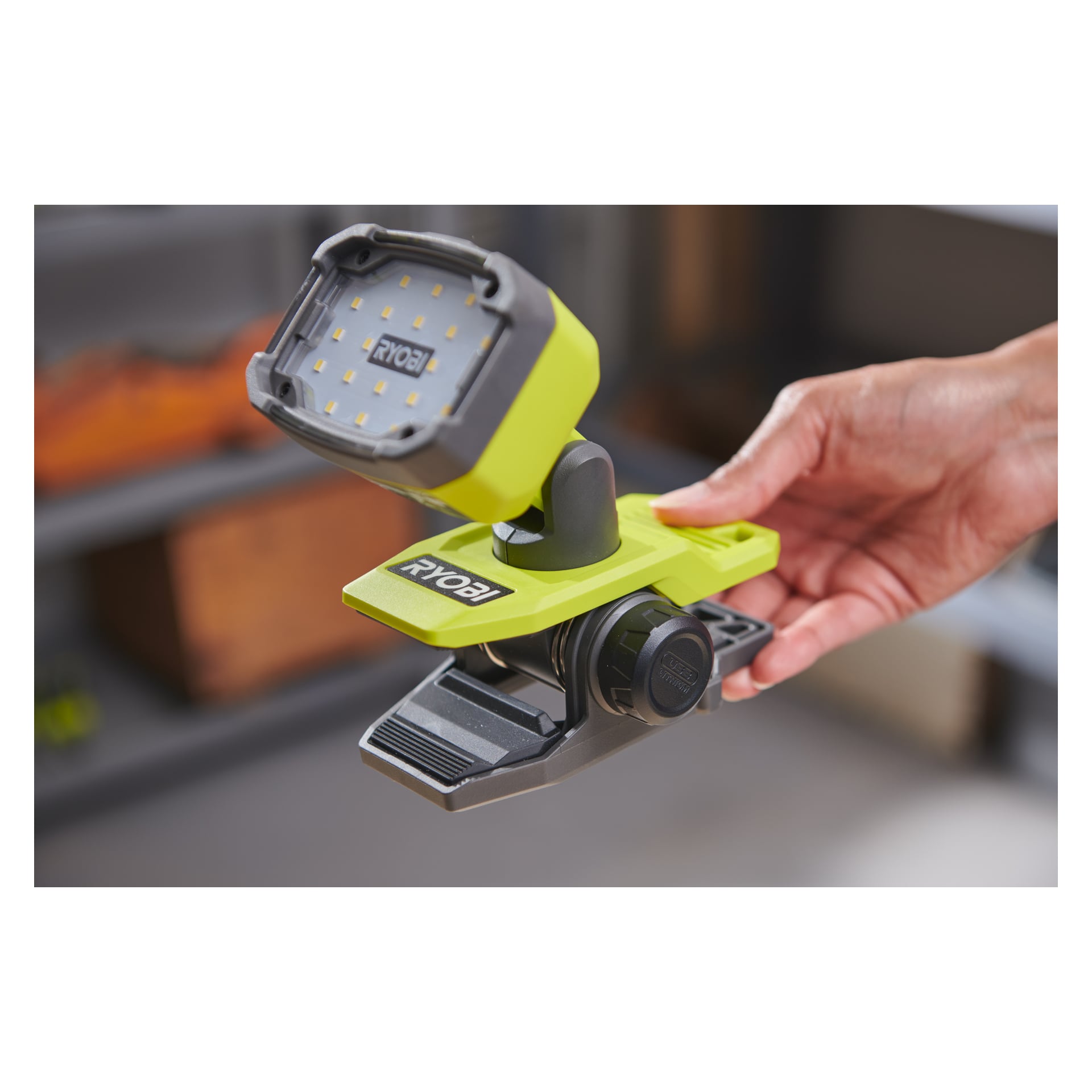 En hand håller en Ryobi LED RLC4-0 arbetslampa, driven av ett USB-litiumbatteri. Lampan har ett grönt och svart plasthölje, ett rektangulärt ljushuvud och en integrerad klämma vid basen för att fästa den på olika ytor.