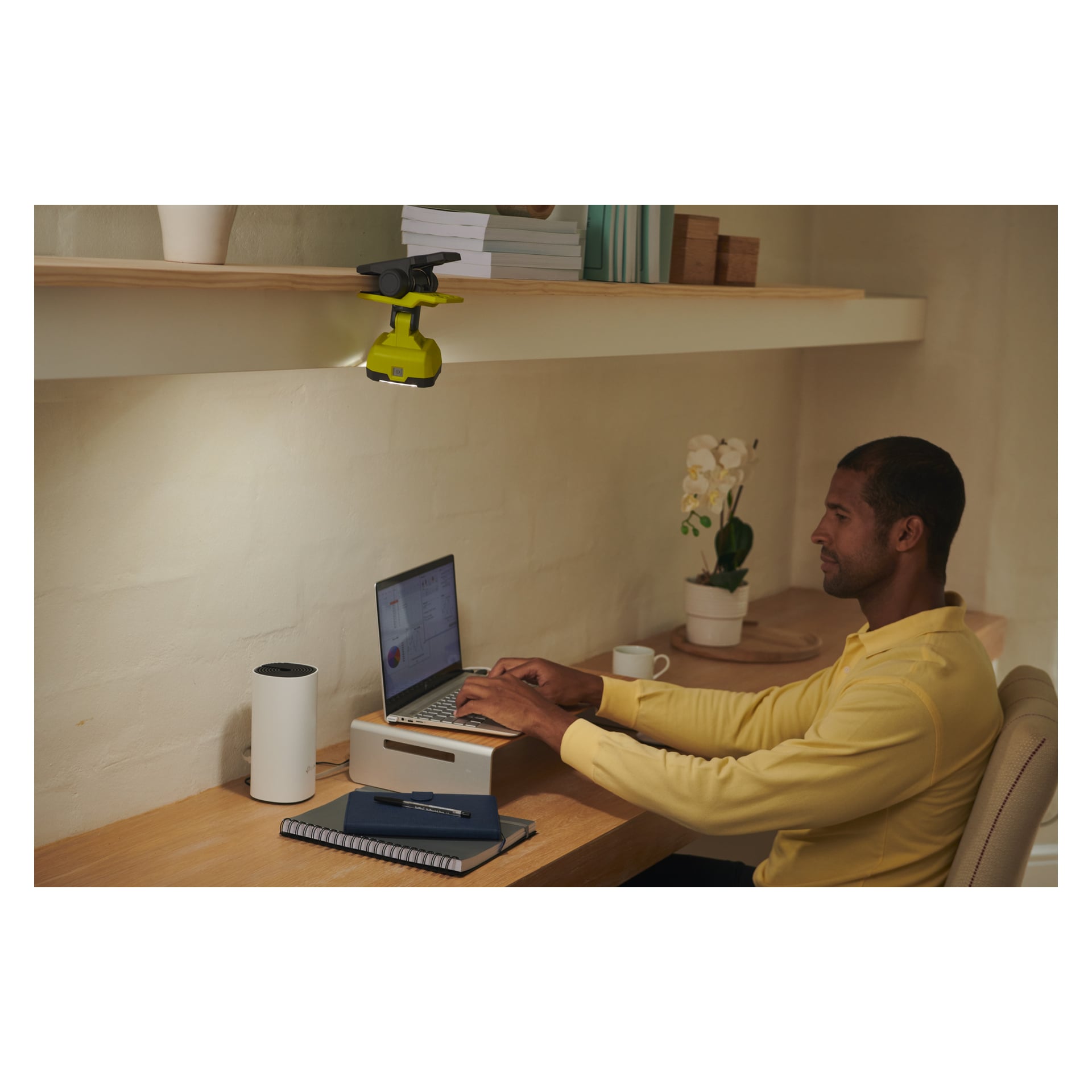 En man i gul skjorta använder en laptop vid sitt hemmakontor, som också har en anteckningsbok, mugg och en liten vit enhet. Ovanför skrivbordet är en Ryobi USB Lithium LED arbetslampa RLC4-0 fäst på hyllan. En orkidéplanta står i bakgrunden.