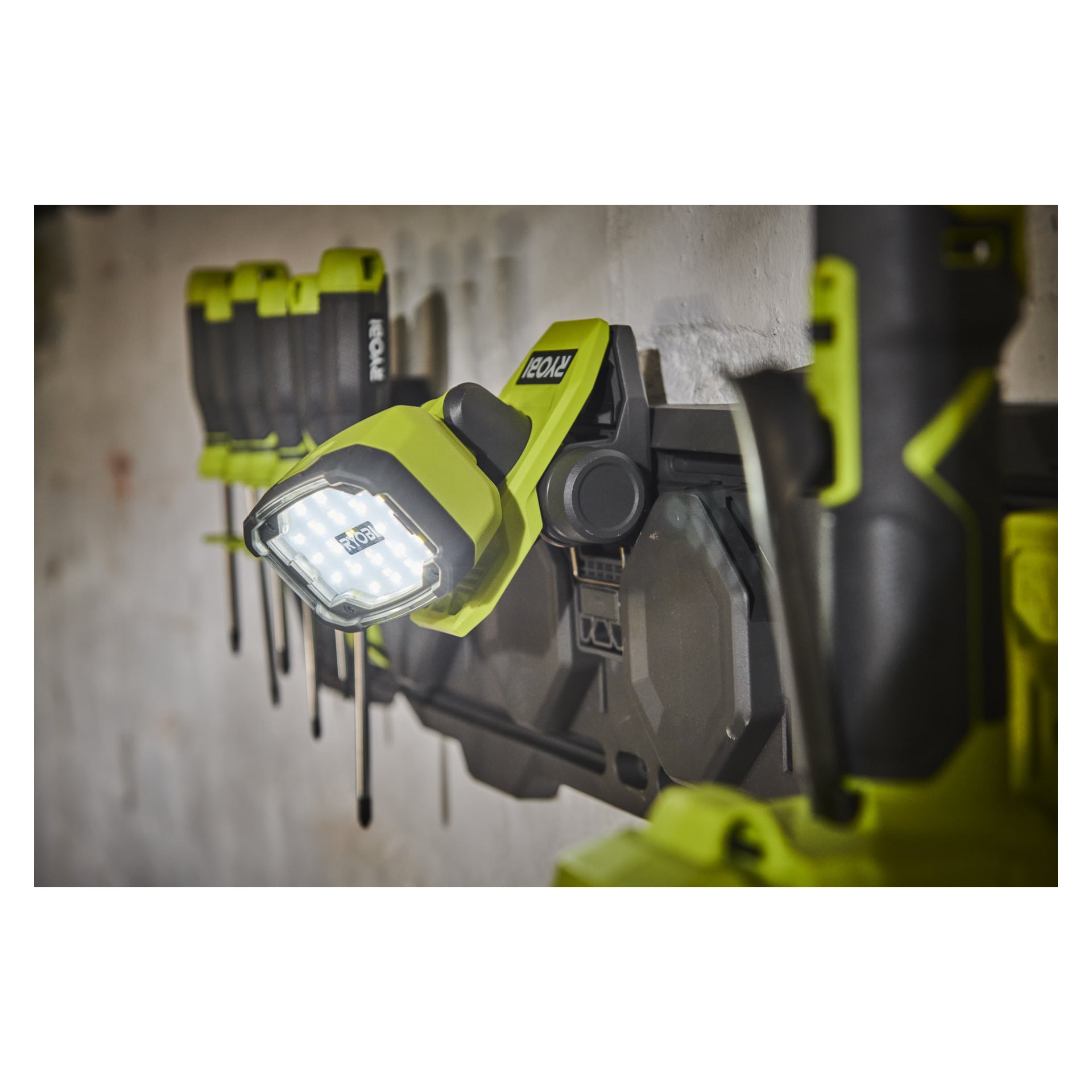 En grön och svart Ryobi USB Lithium Battery-driven LED-arbetslampa (RLC4-0) är fäst vid ett grått förvaringsställ bland olika andra organiserade Ryobi-verktyg, alla säkert placerade på en vit vägg.