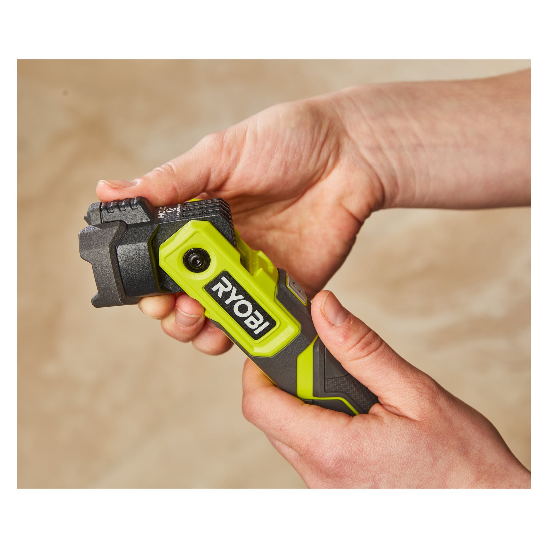 Någon håller en grön och svart Ryobi USB Lithium Rechargeable LED-ficklampa RLP4-0 med båda händerna och vrider på en ratt eller strömbrytare på toppen. Bakgrunden är enkel och ljusbrun.