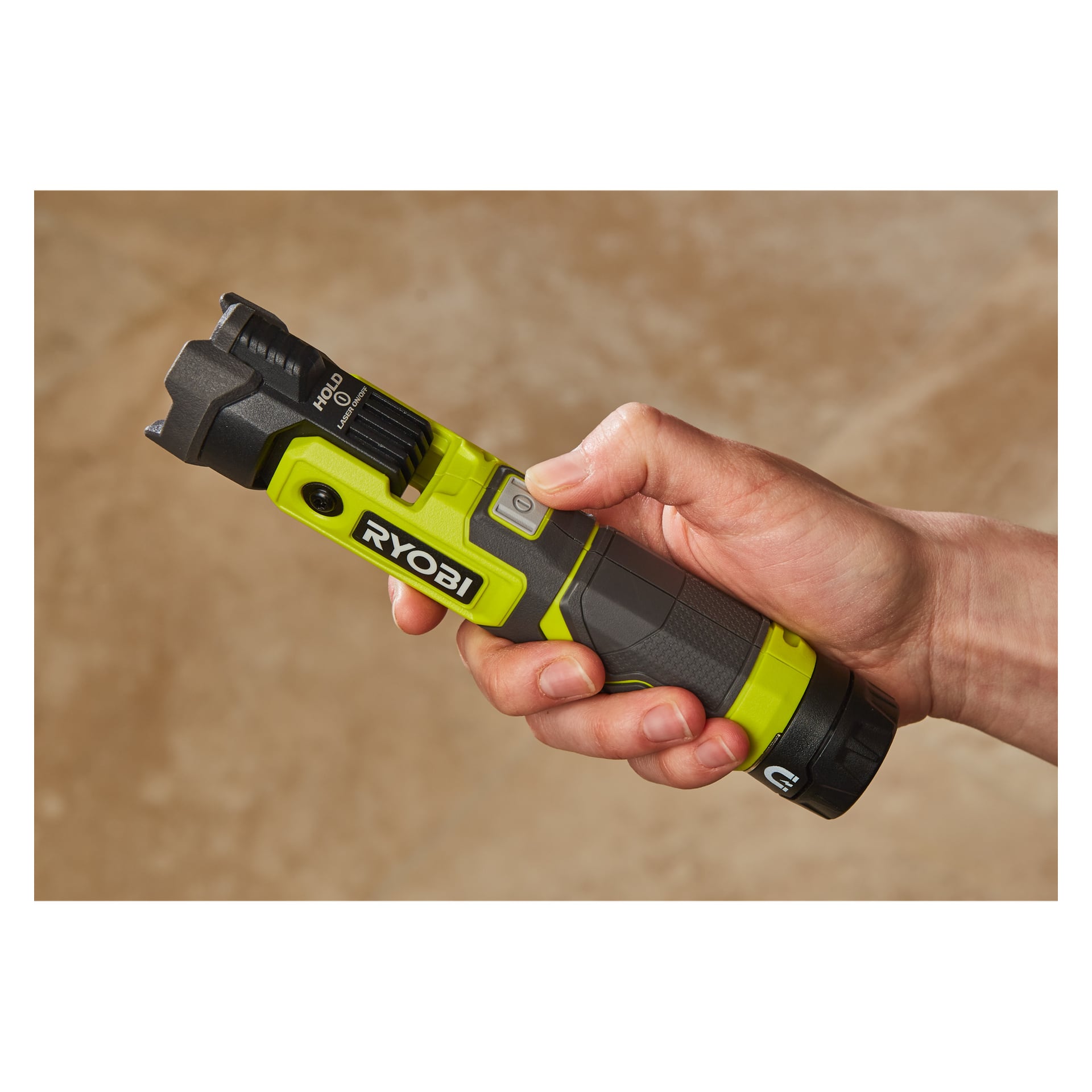 En hand håller en grön och svart Ryobi USB Lithium Rechargeable LED-ficklampa RLP4-0 med en synlig strömbrytare och en delvis synlig hålletikett nära toppen. Bakgrunden är en neutral brun yta.