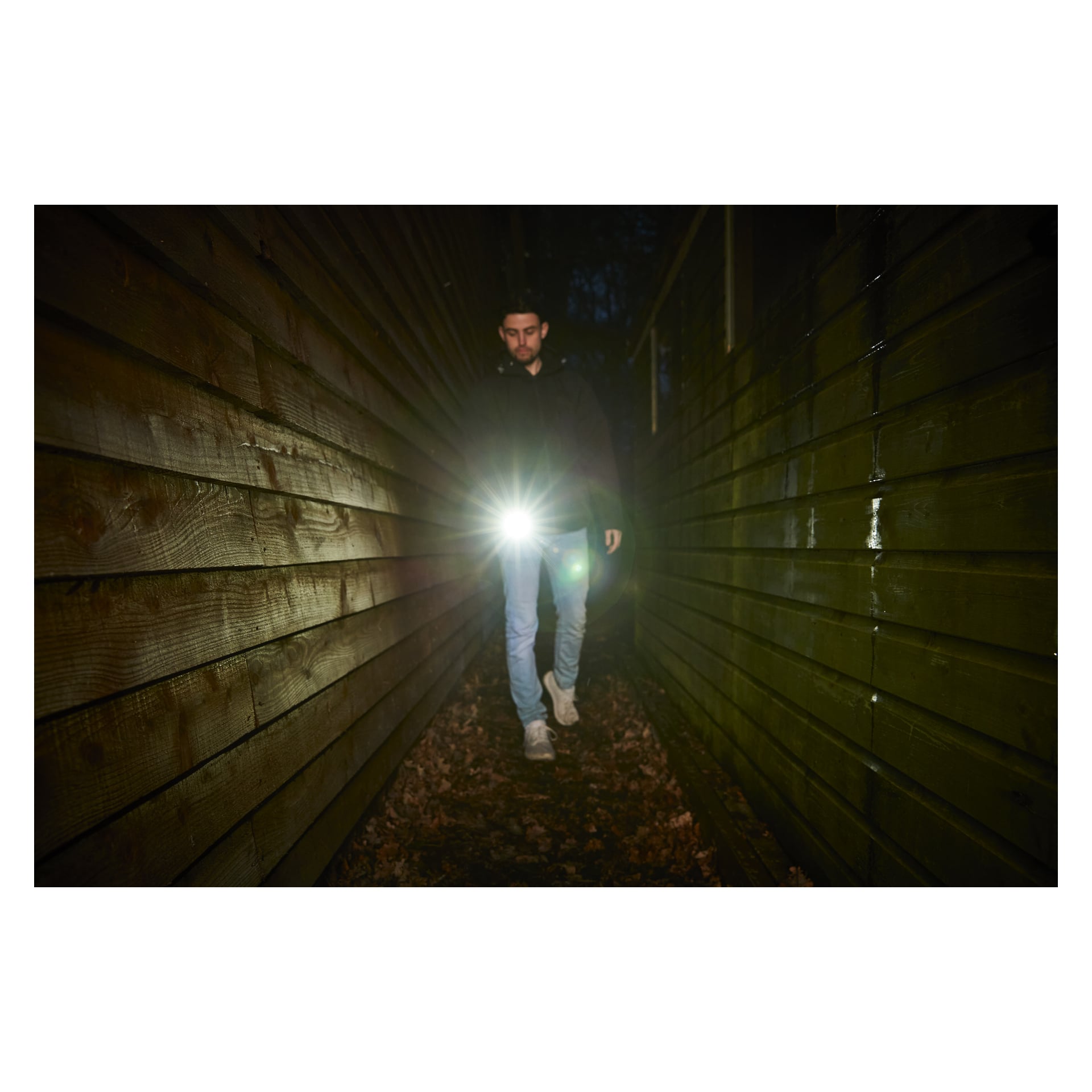 En person går på natten mellan två träväggar och lyser upp marken täckt av utspridda löv medan de lyser med en Ryobi USB Lithium Rechargeable LED-ficklampa RLP4-0 mot kameran.