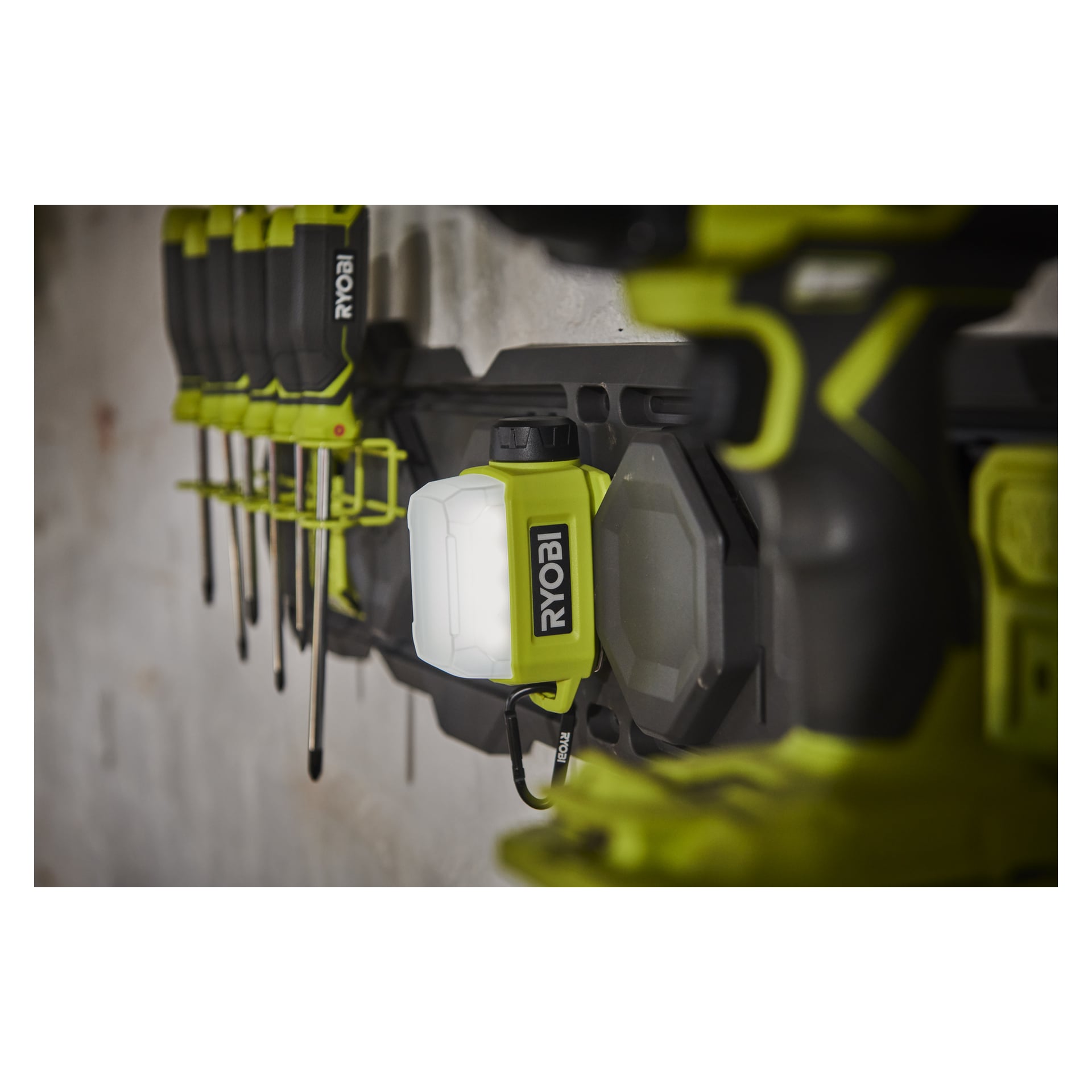 En Ryobi USB Lithium Battery-driven områdeslampa RLA4-0 är monterad på ett väggställ bredvid andra Ryobi-verktyg och skruvmejslare i en verkstad, med väggen bakom dem som ser ofärdig eller grovmålad ut.