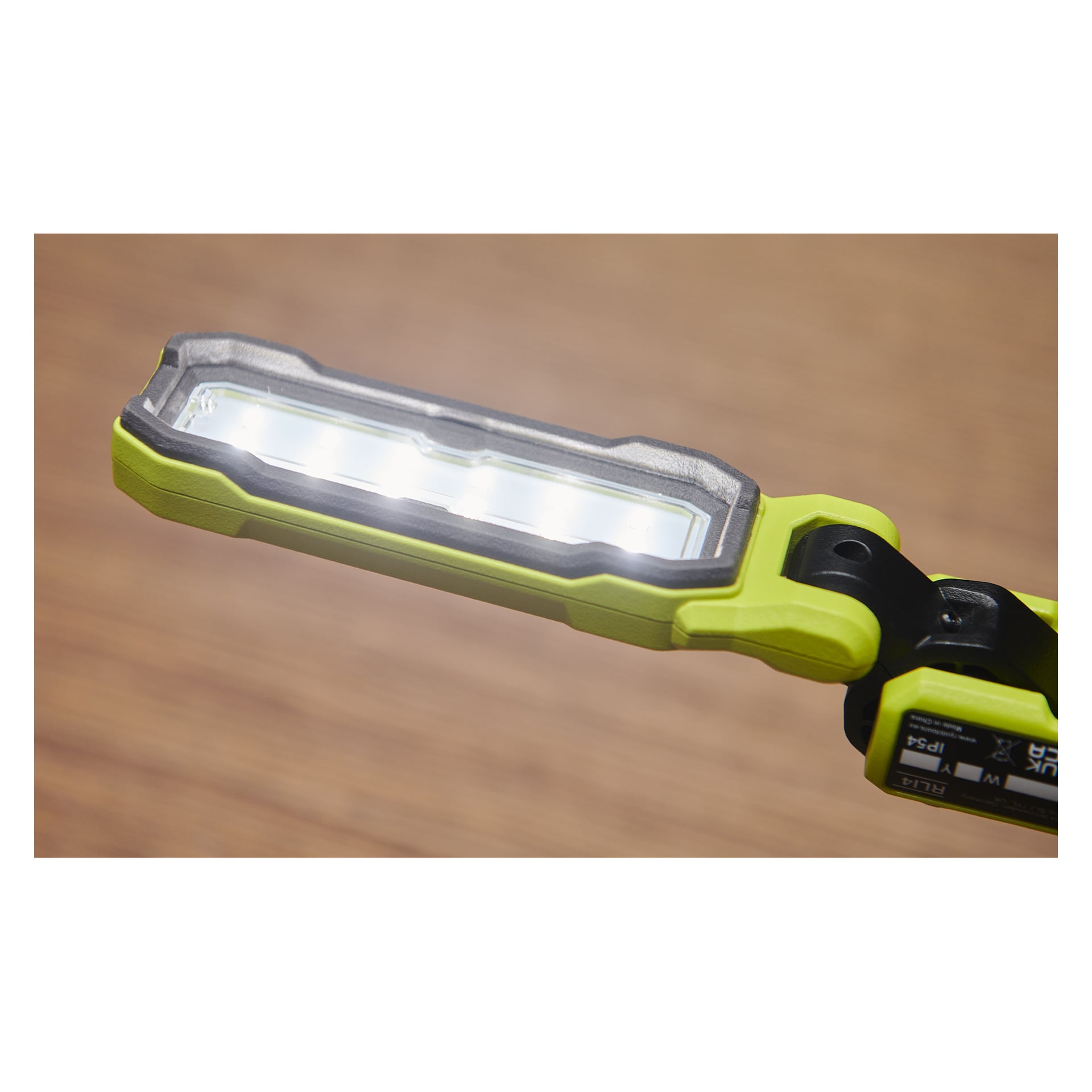 En närbild av Ryobi USB Lithium Work-lampan RLI4-0, en rektangulär LED-lampa i grönt och svart, tänd och lysande starkt vitt ljus mot en mjukt suddig brun bakgrund.